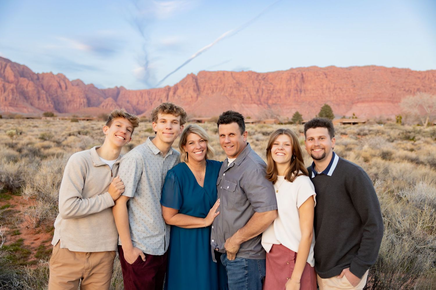 A family of 6 is standing closely together in front of the beautiful Southern Utah red mountains.