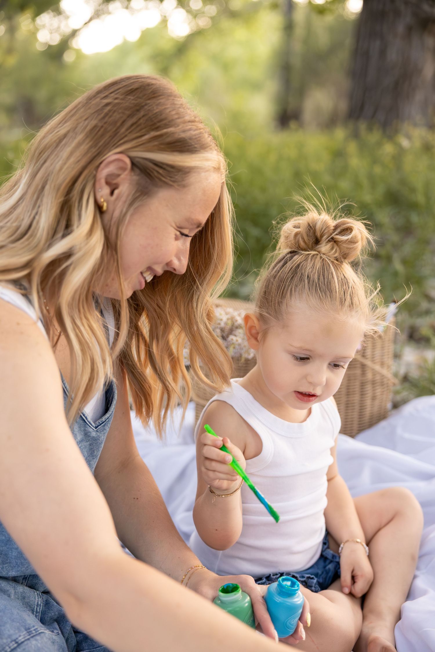A mom and her daughter are sitting in a field painting a canvas together.