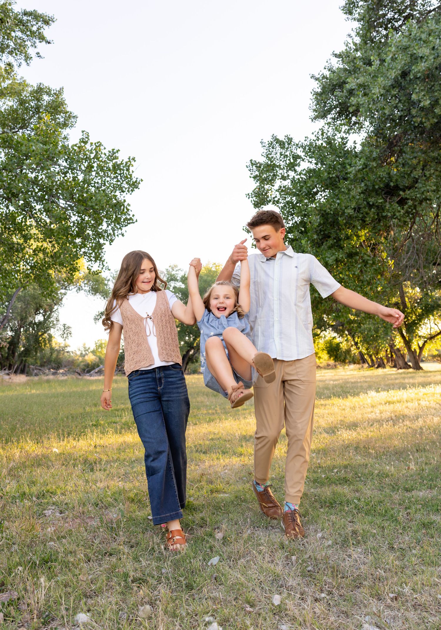 3 siblings are holding hands. The youngest in the middle is getting swung in the air by the older two, while they all giggle at each other.