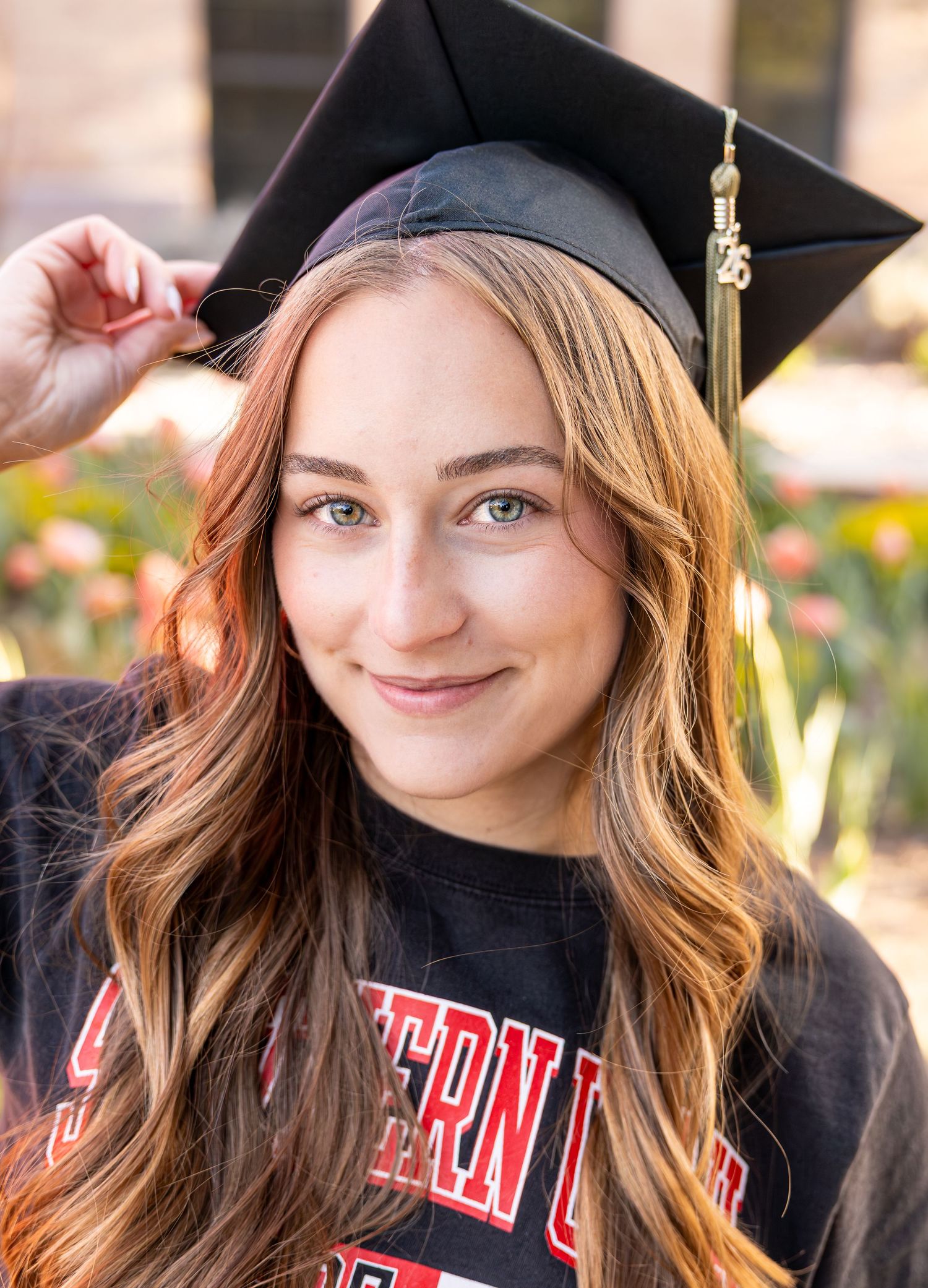 A college graduate is sitting in front of a field of flowers with her cap and gown on.