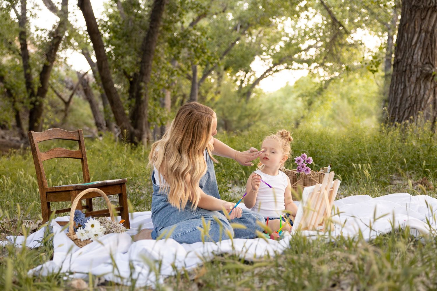 A mom and her daughter are sitting in a field painting together on a canvas and on each other.