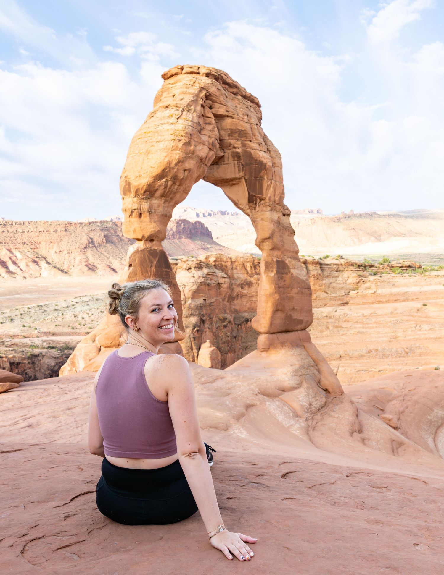 A girl is sitting on a rock in front of Delicate Arch in Moab Utah.