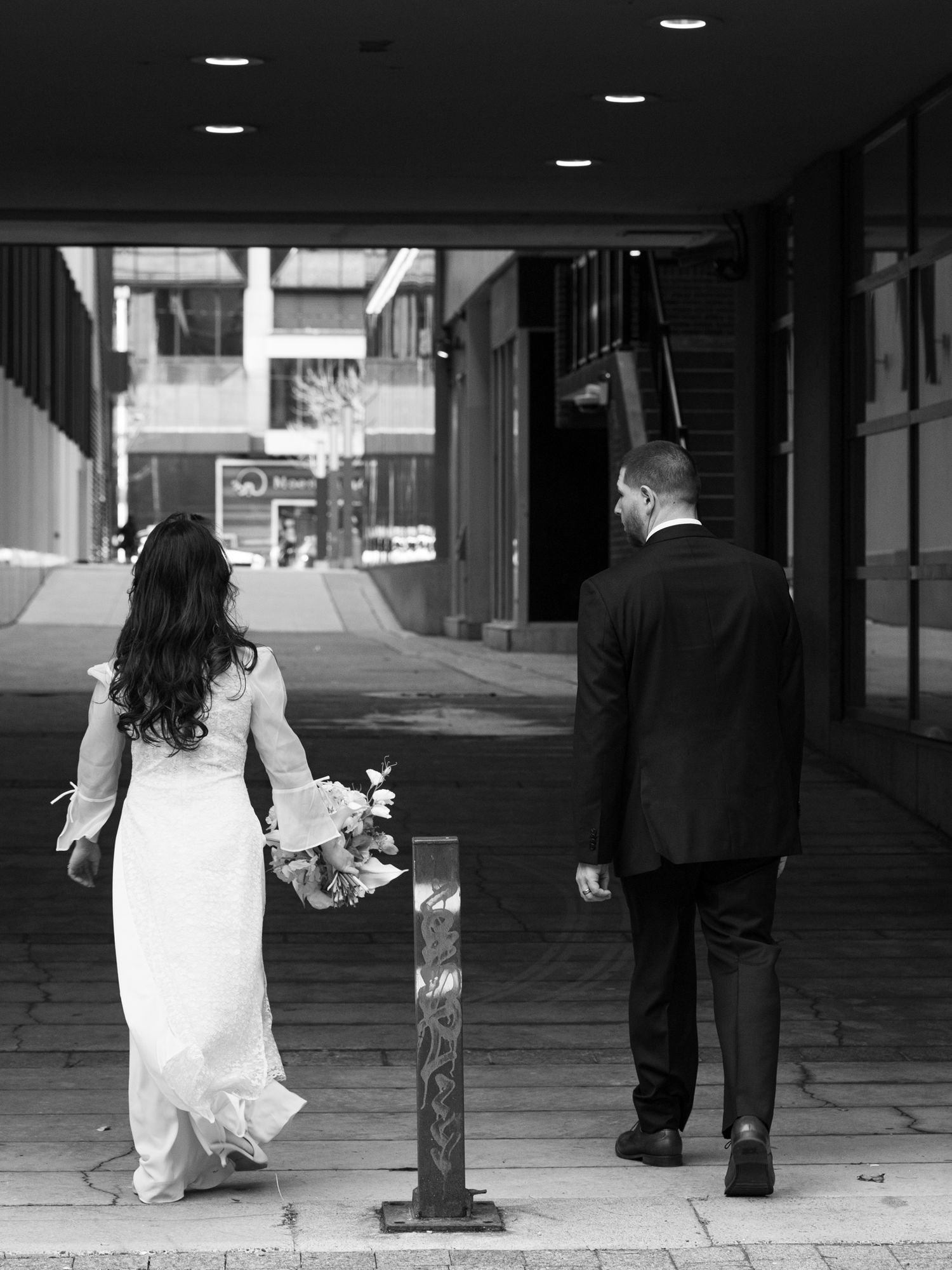 Black and white photo of a bride and groom walking away down a city sidewalk, bride holding a bouquet.