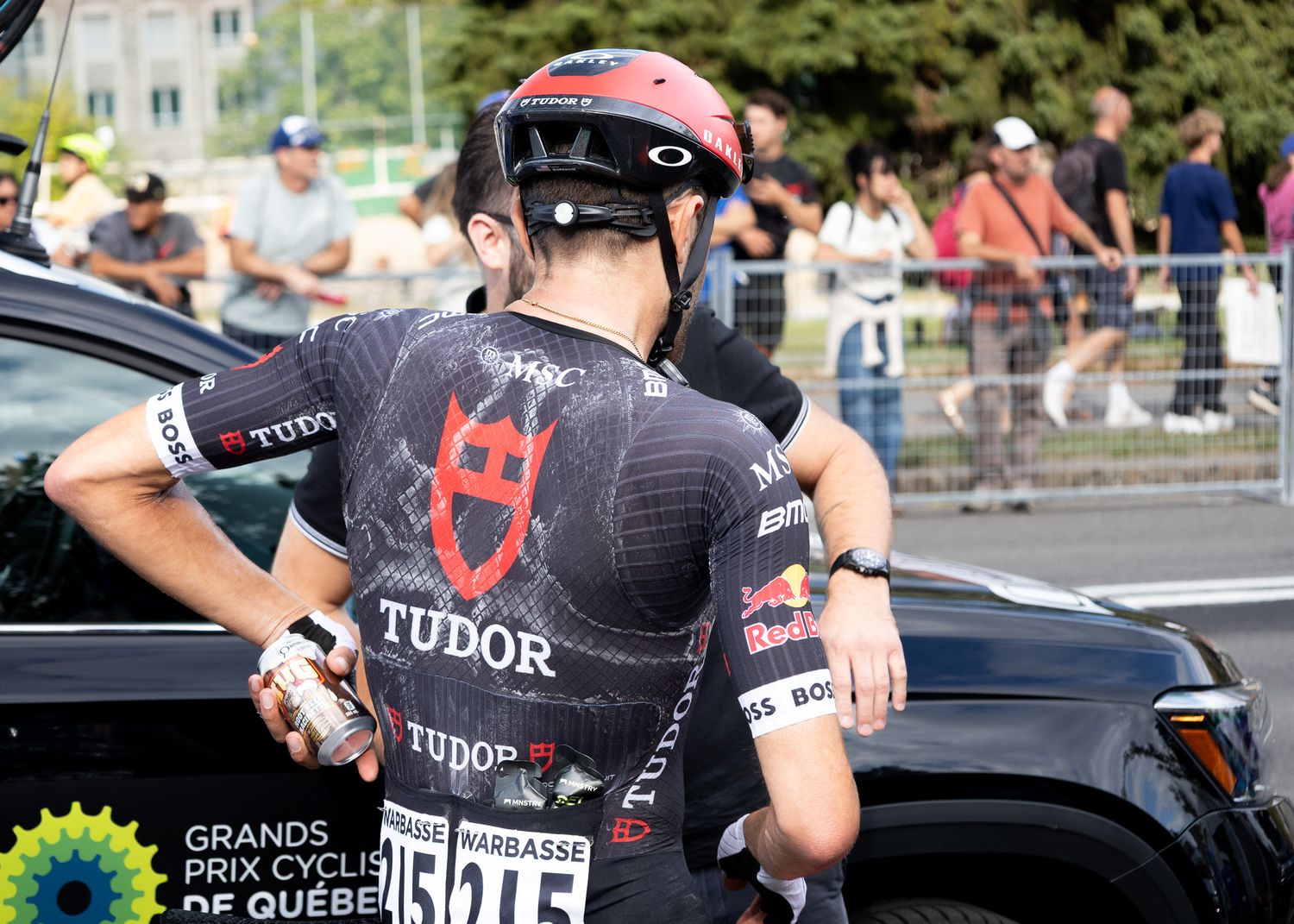 Professional cyclist in Tudor team jersey waits near support vehicle during cycling race event.