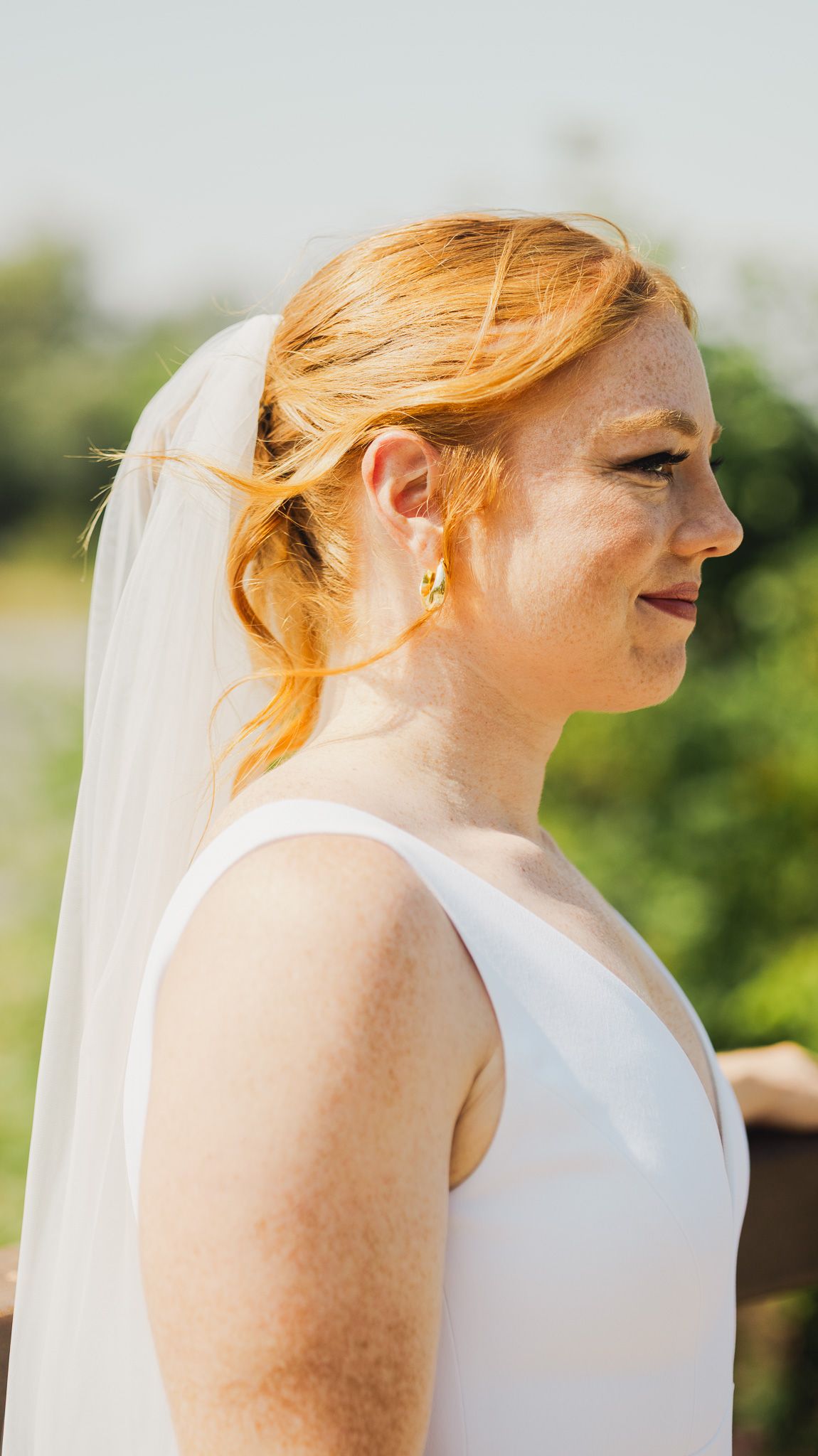 Bride wearing a white sleeveless dress with flowing veil against a natural outdoor backdrop.
