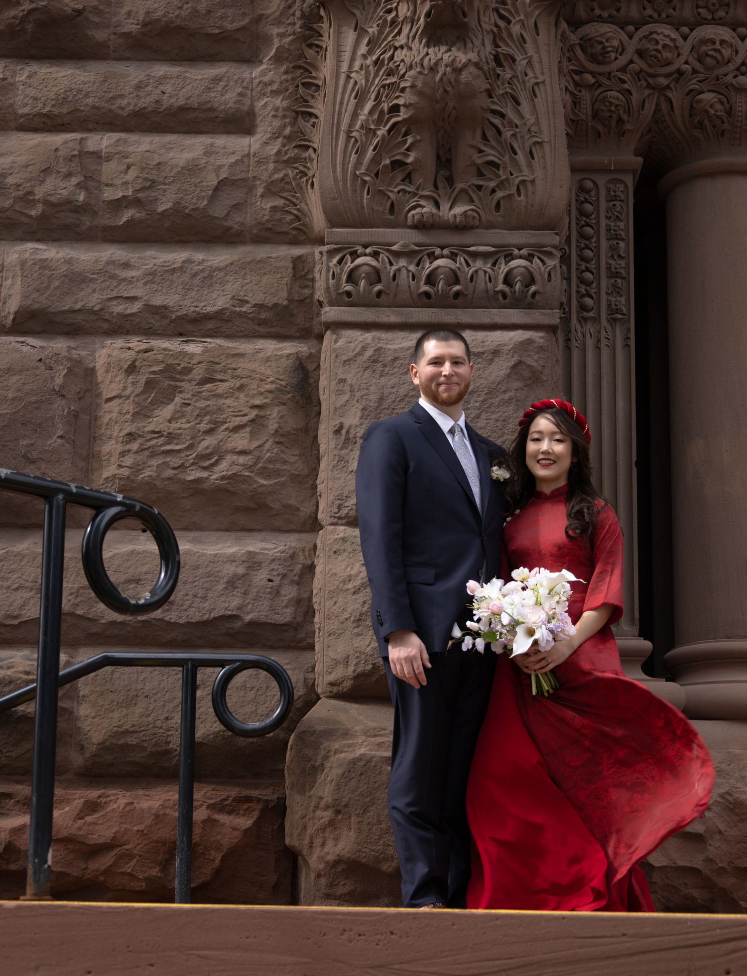 Bride in red dress with floral crown holds white bouquet beside groom in navy suit on church steps.