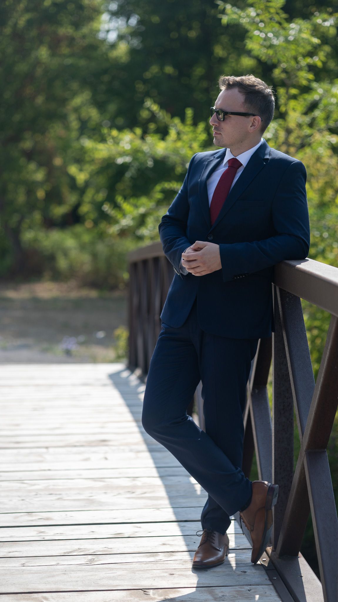 Groom in navy suit sitting casually on wooden deck railing.