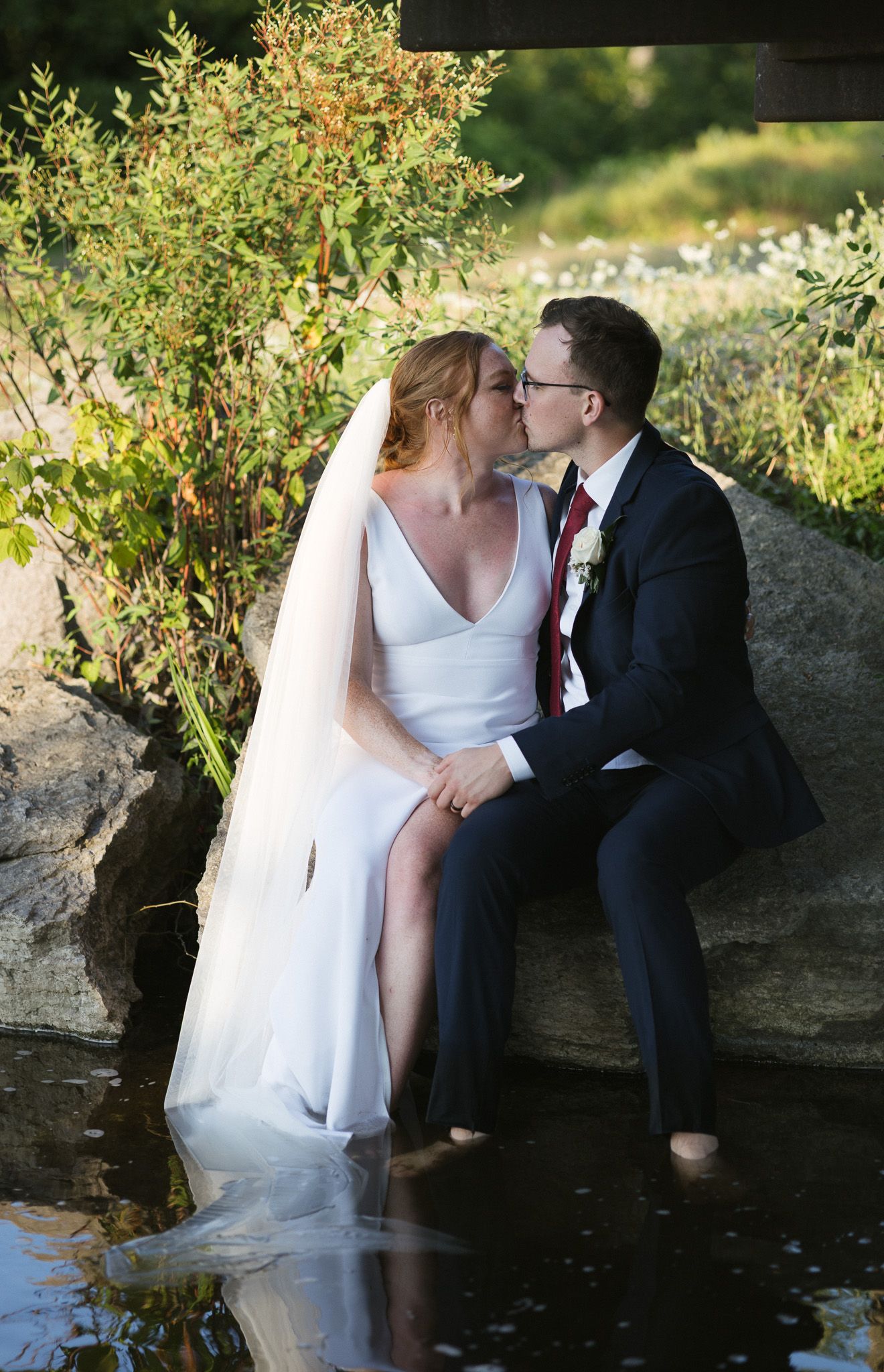 Newlyweds sharing intimate moment by water's edge with greenery in background.
