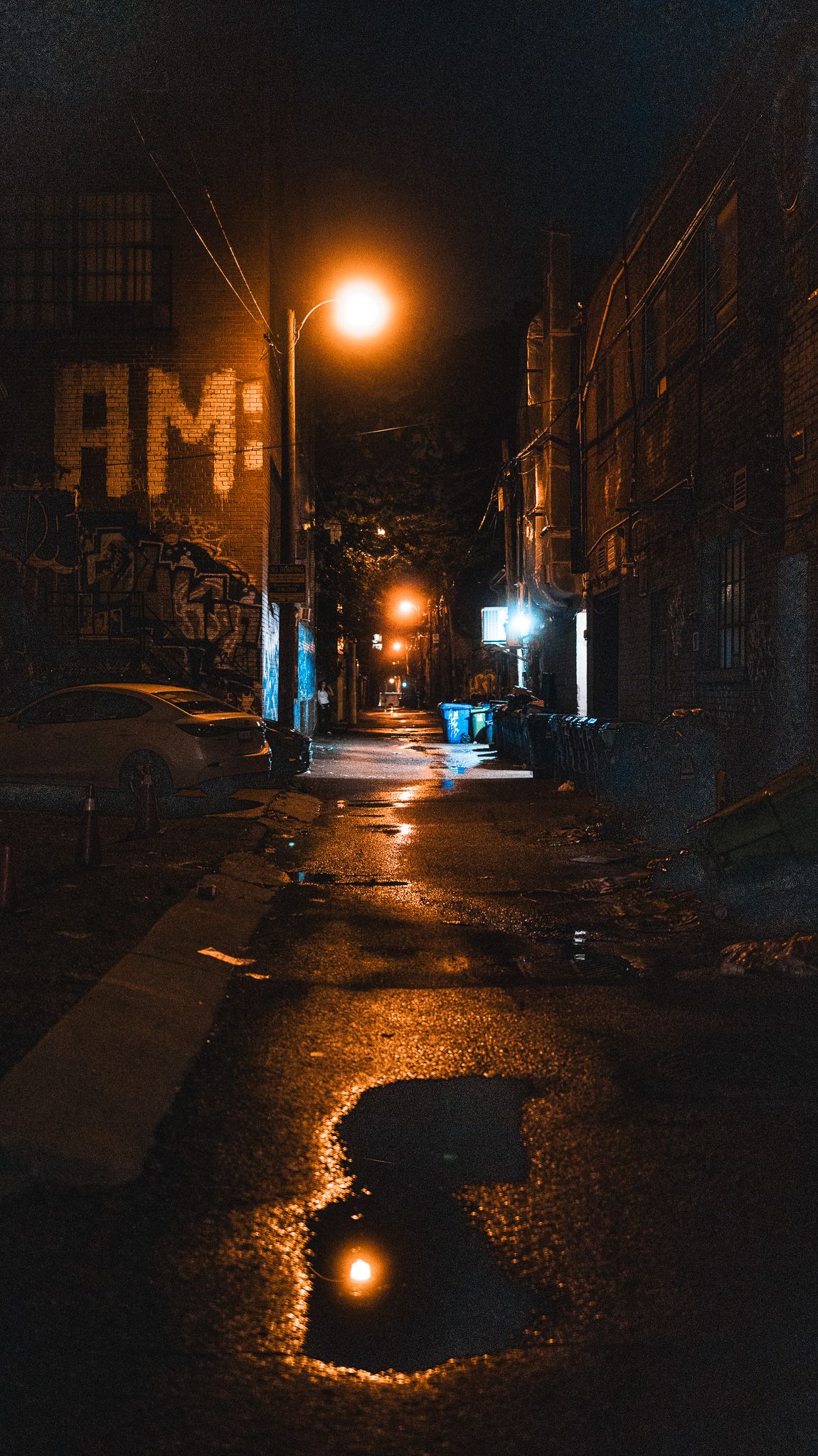 Dark urban street at night with glowing orange street lamps reflecting on wet pavement.
