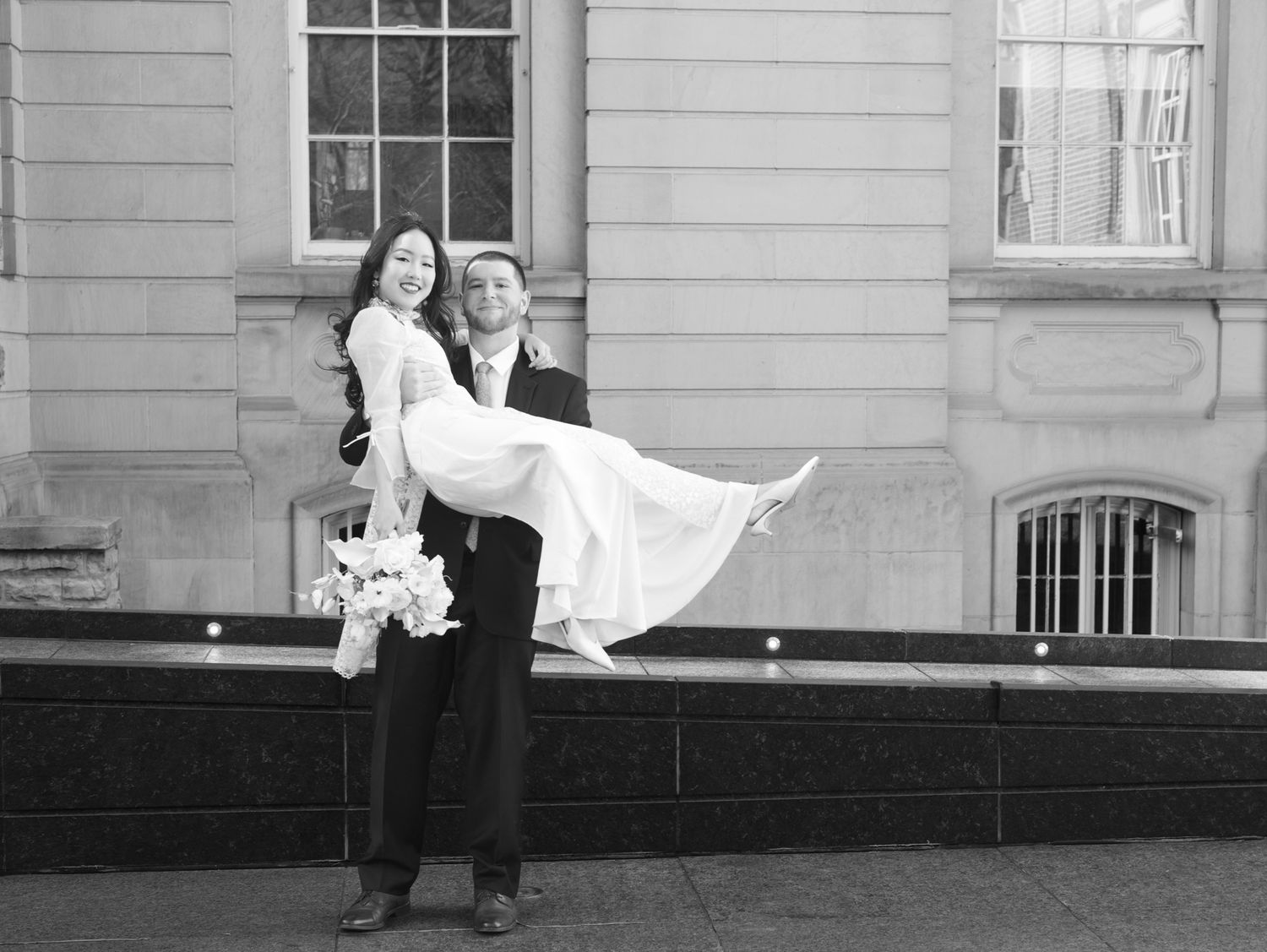 Groom carries bride in white dress outside historic building in classic black and white wedding photo.