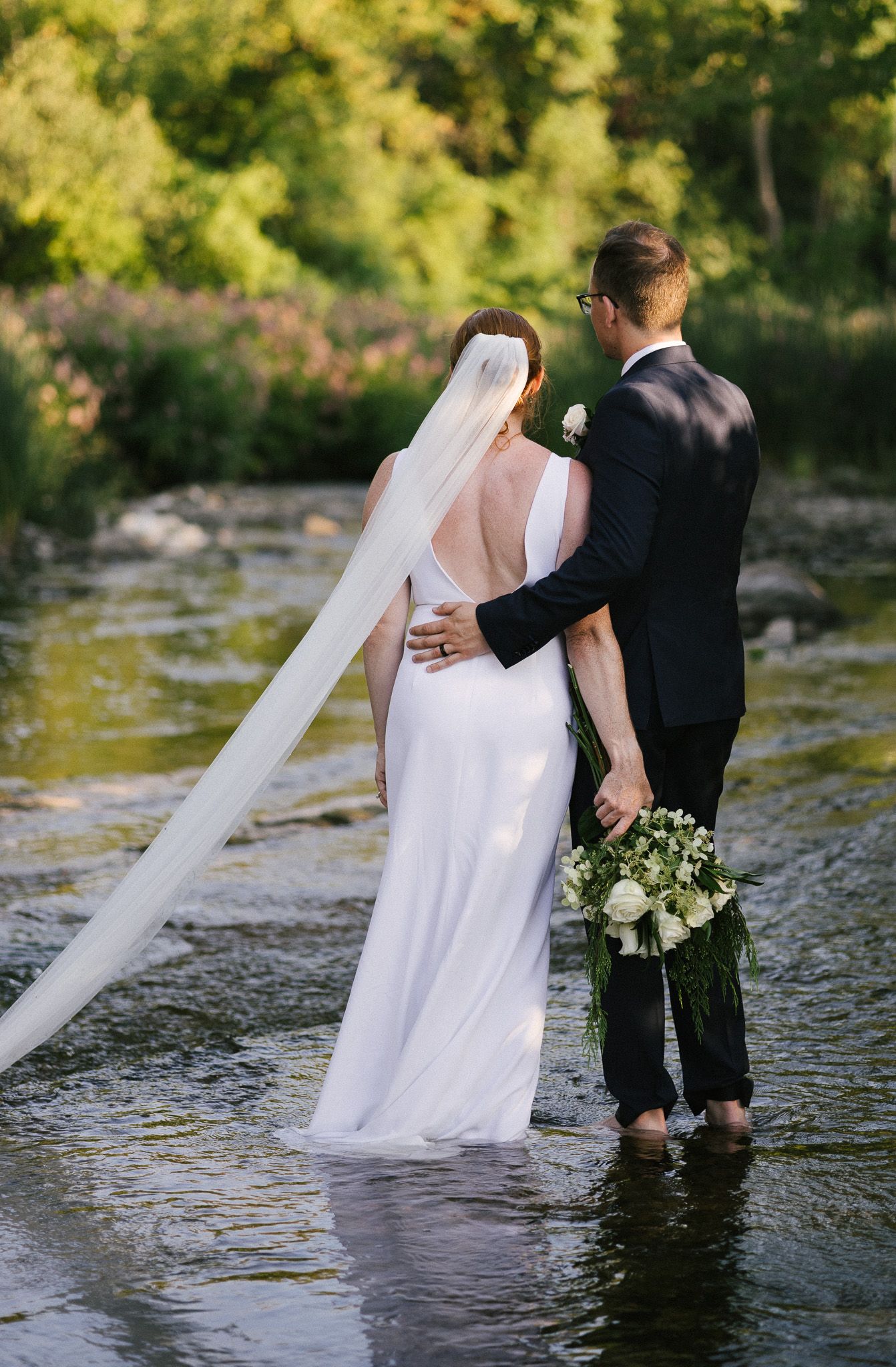 Wedding couple standing in shallow river water with flowing white dress and bouquet in scenic outdoor setting.