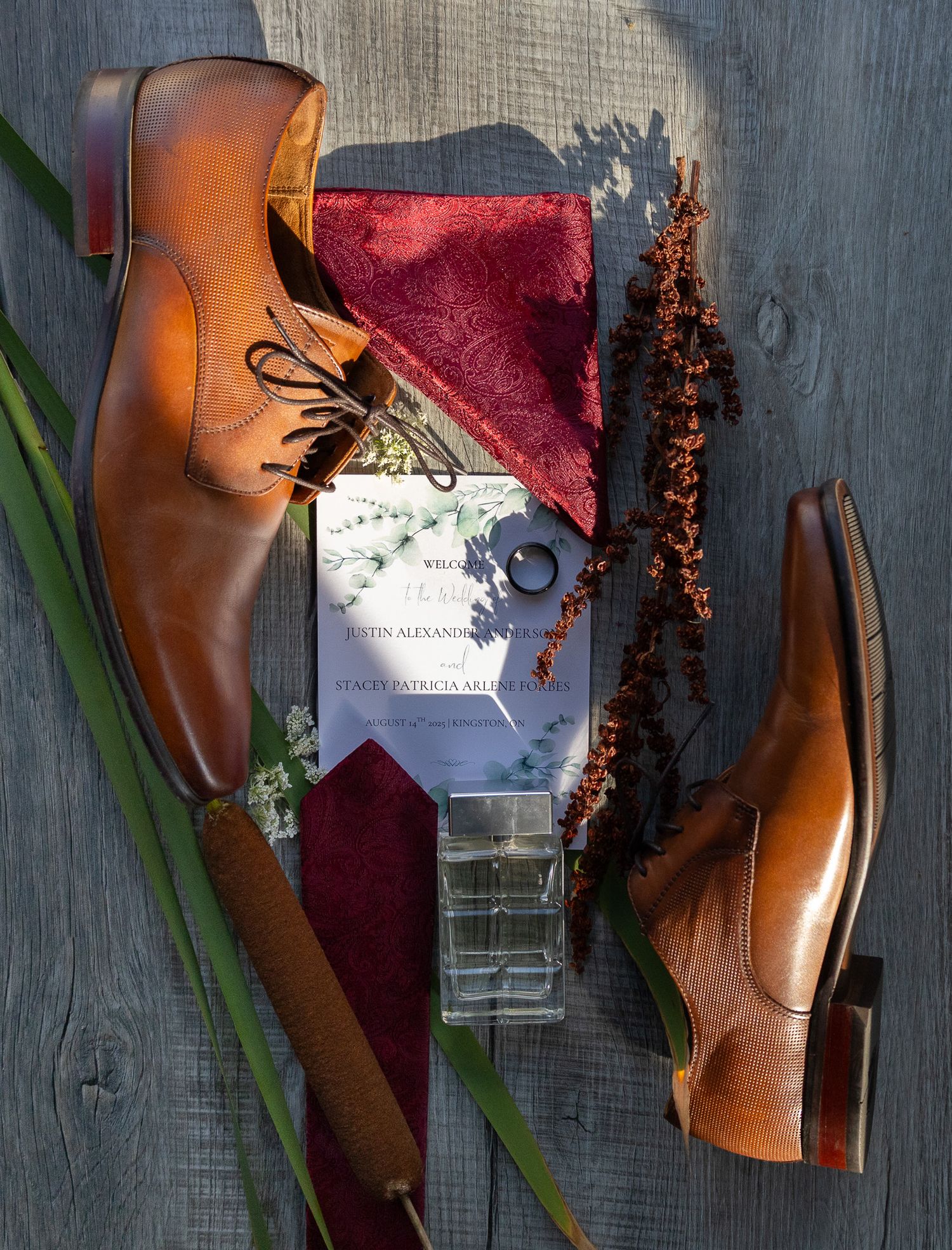 Brown leather dress shoes with burgundy accessories and wedding invitation on gray wood background.