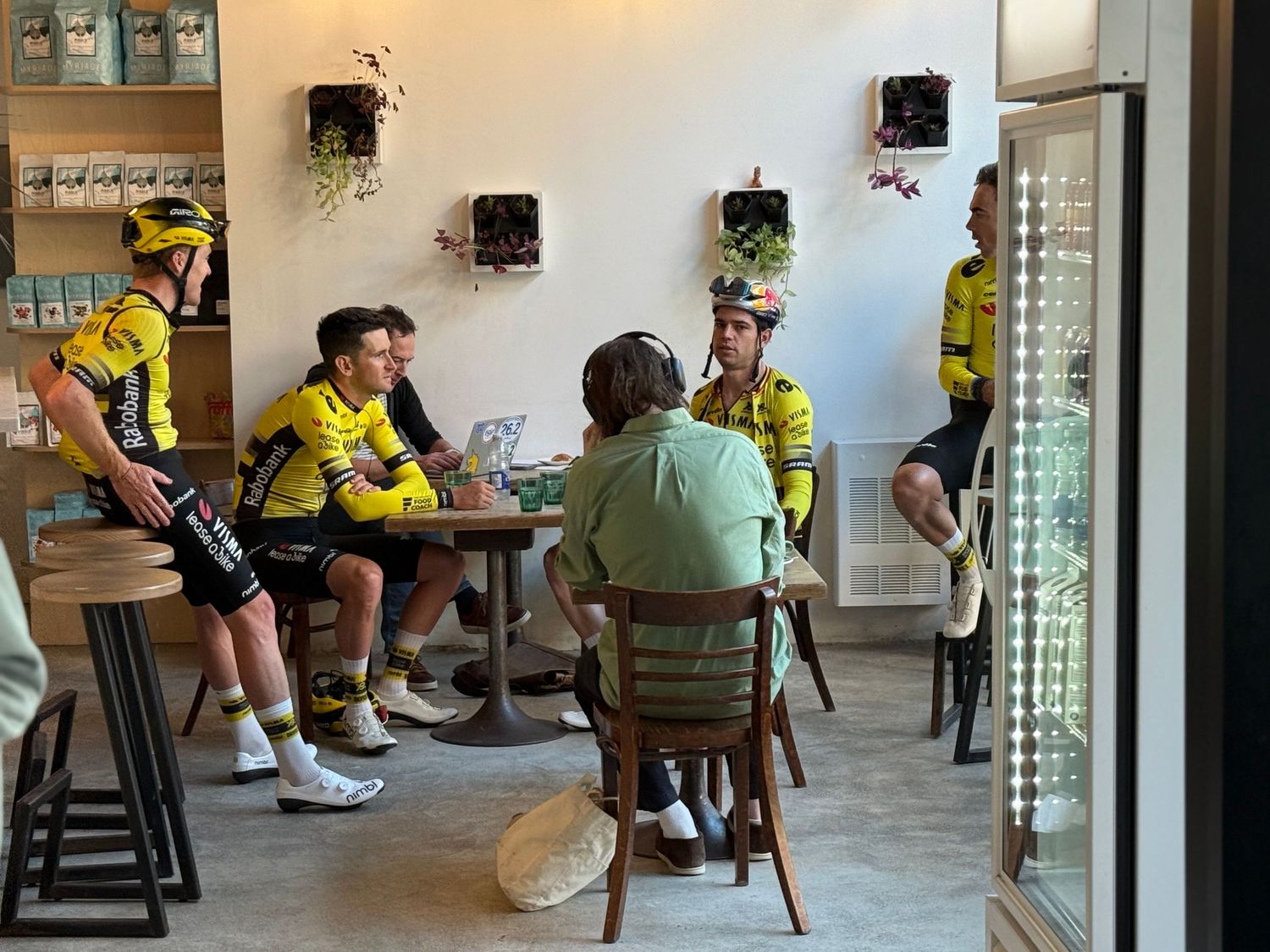 Cyclists in yellow jerseys relax at a cafe counter with stools and glass block windows.