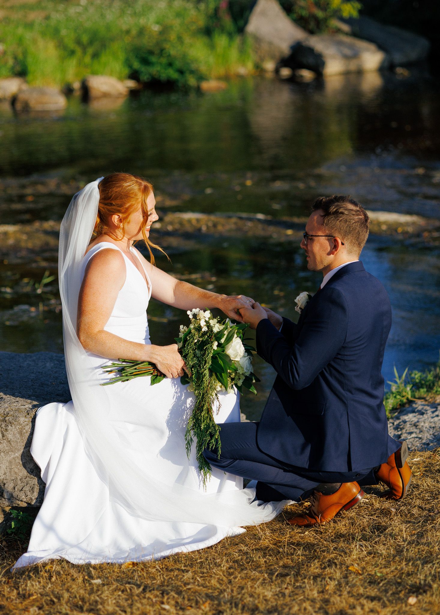 Newlywed couple sits together by a tranquil pond on a sunny afternoon with greenery in the background.