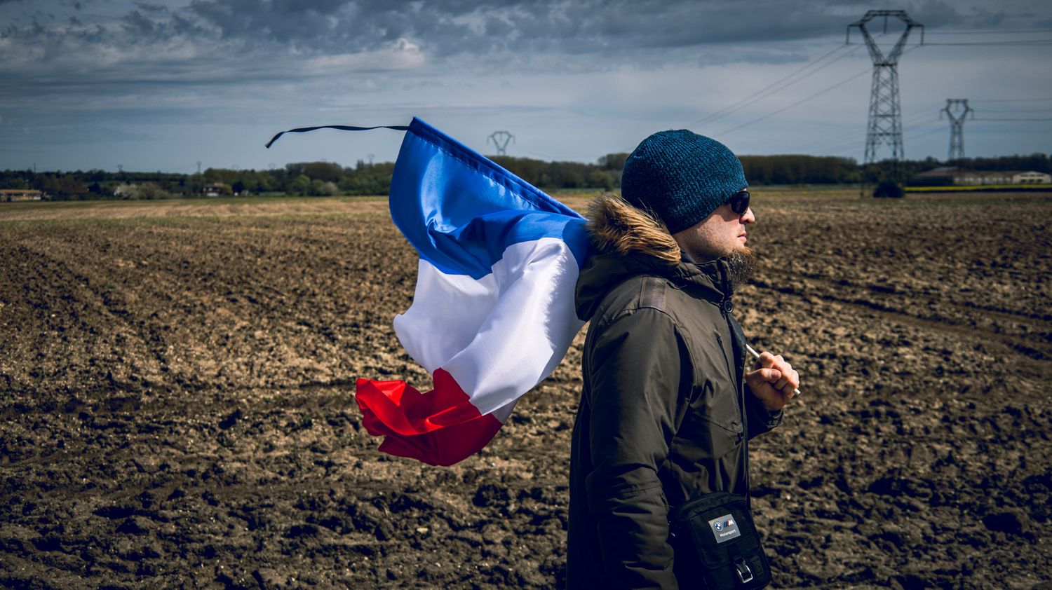 French flag waves in wind against cloudy rural landscape with power lines.