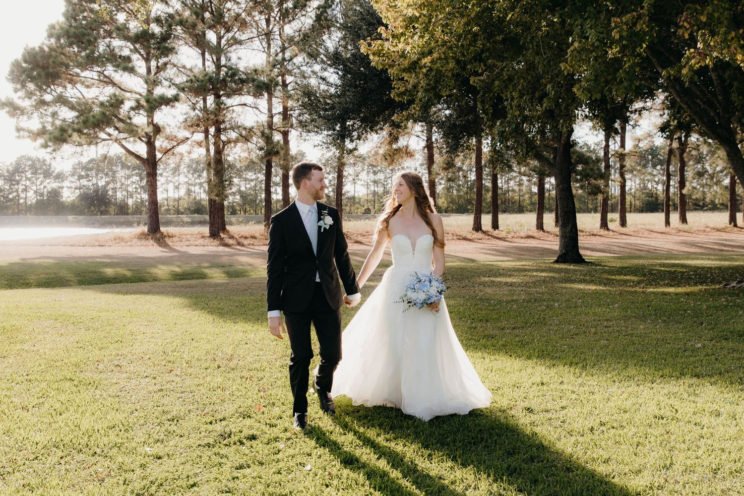 A couple in wedding attire walks together on a sunlit grassy lawn with trees in the background.