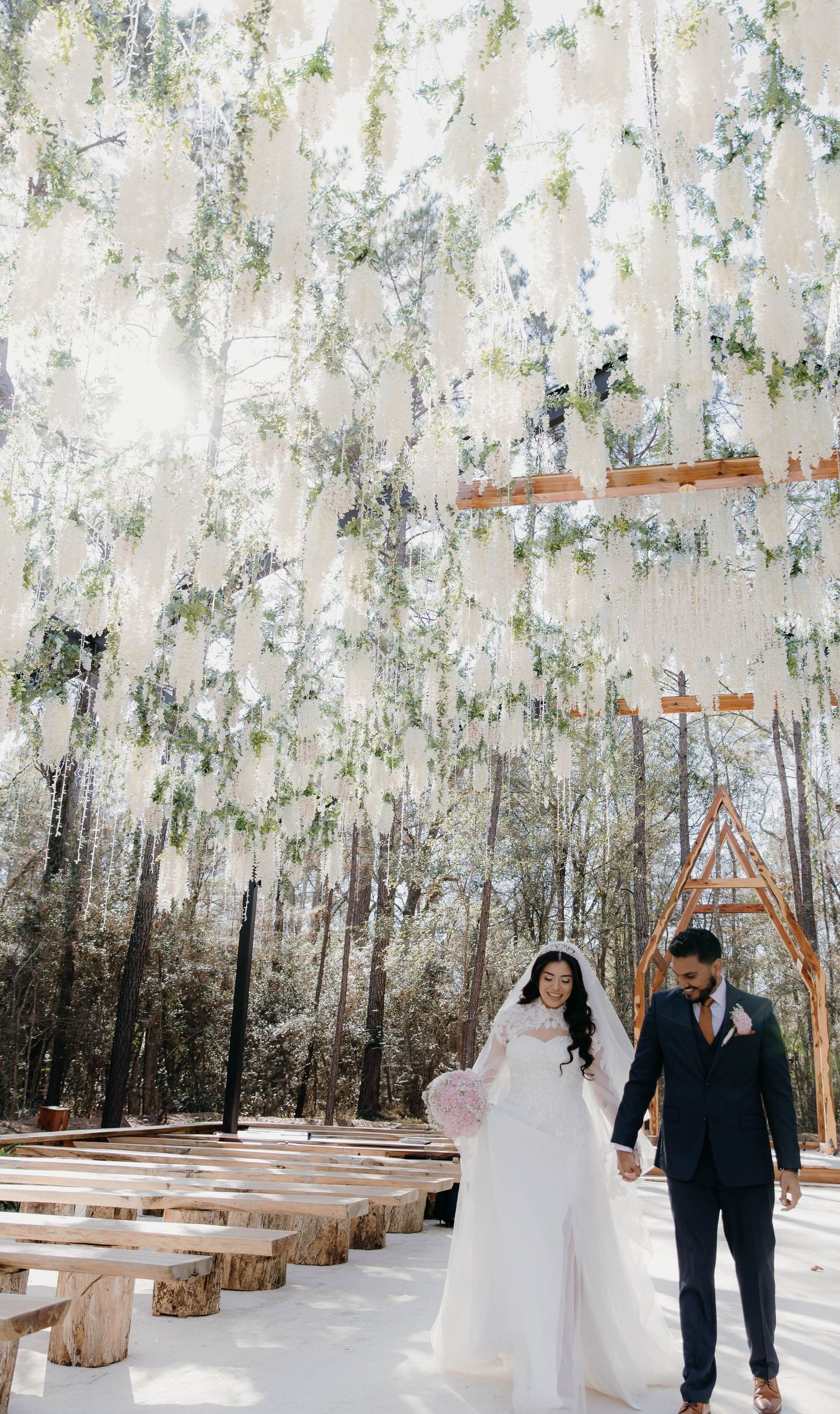 Couple walking through an enchanted forest setting with white floral decor and wooden benches for an outdoor wedding ceremony.