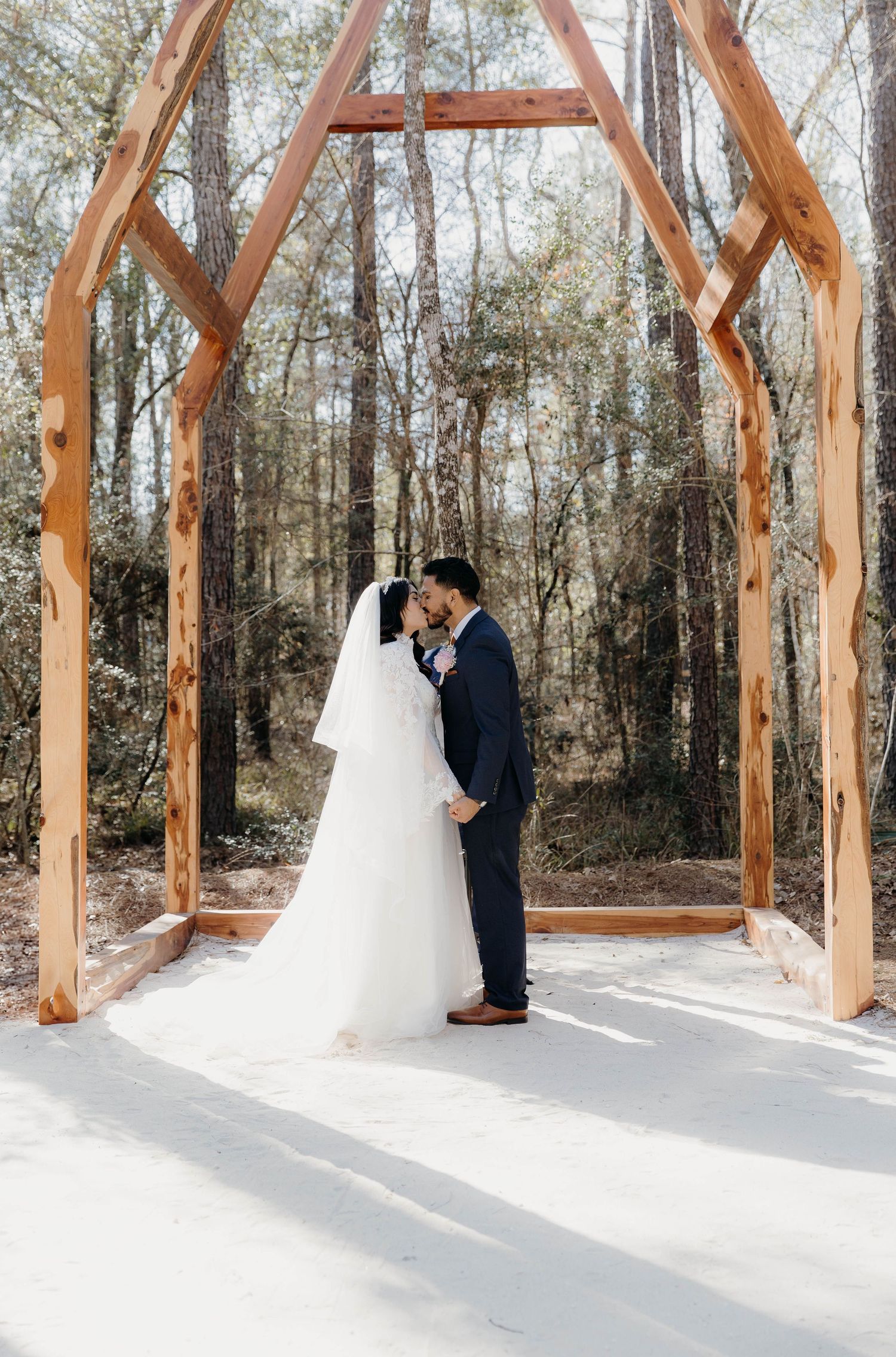 A romantic wedding couple embraces under a rustic wooden arch in a forest setting with natural sunlight streaming through.