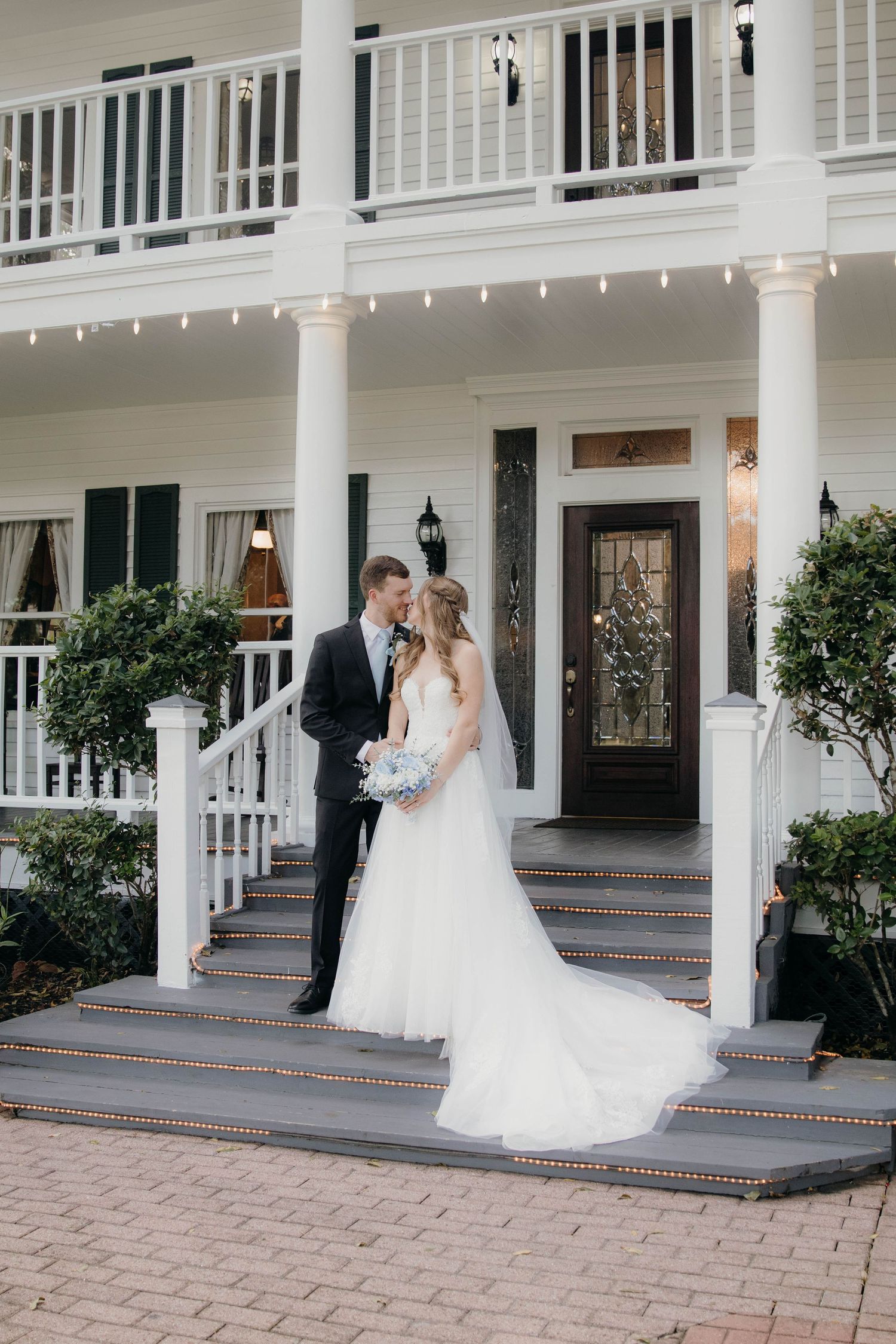 A couple in wedding attire dances together on the porch of an elegant white colonial-style mansion with columns.
