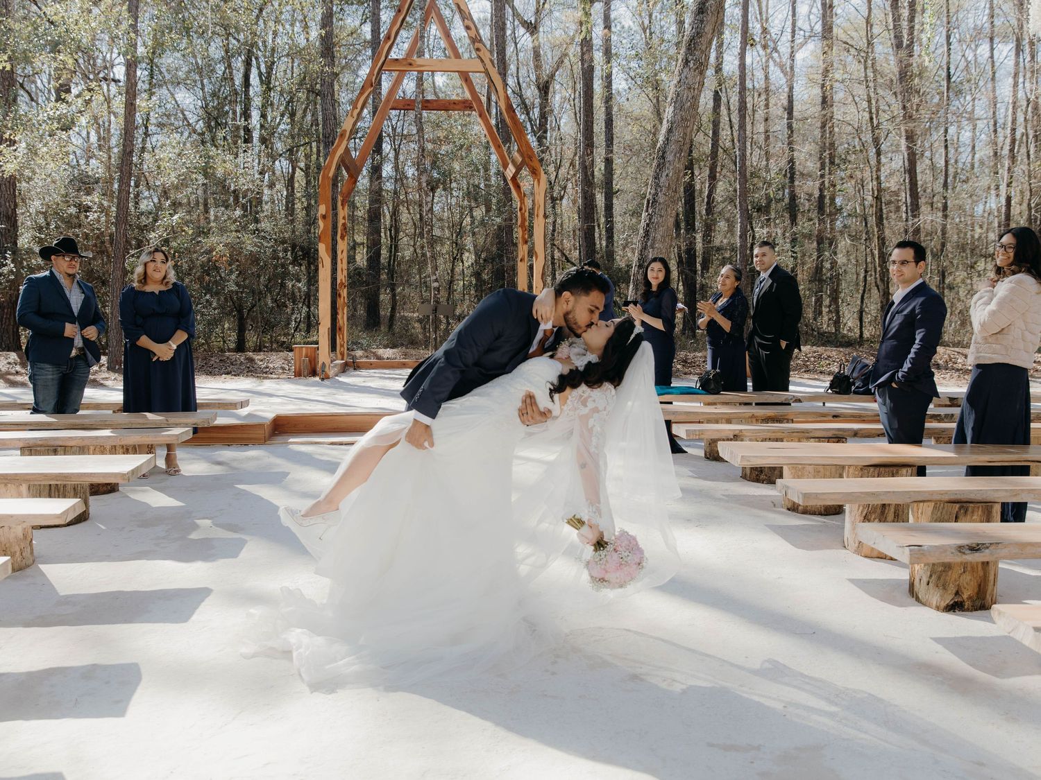 A snowy winter wedding ceremony takes place outdoors under a wooden arch surrounded by tall pine trees.