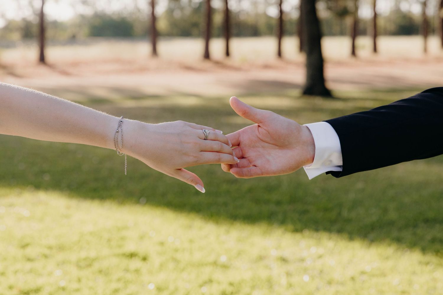 Two hands reach toward each other in the sunlight during an outdoor wedding ceremony.