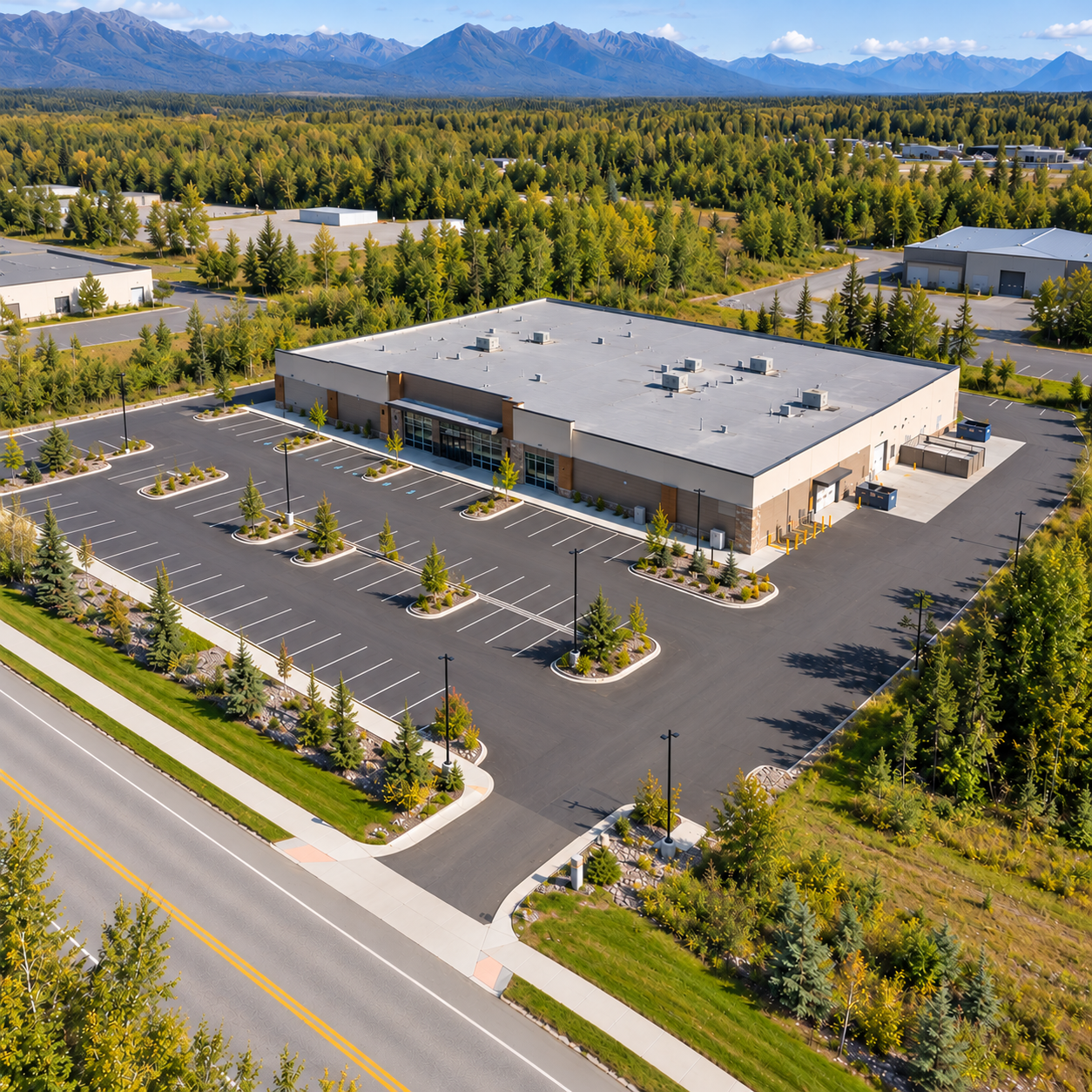 Aerial view of a commercial building with an empty parking lot and landscaped grounds, surrounded by forest with mountains in the distance.