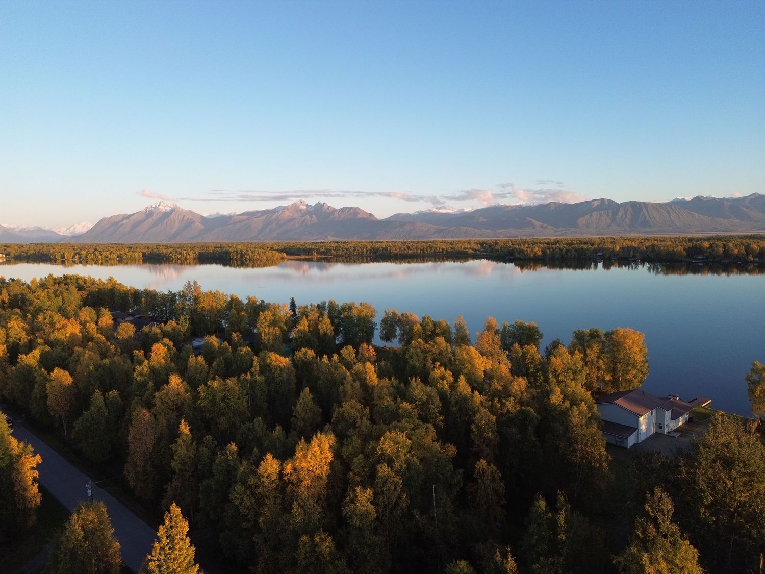 Aerial view of a lake bordered by golden autumn trees and lakeside homes, with snow-dusted mountains beneath a clear blue sky.