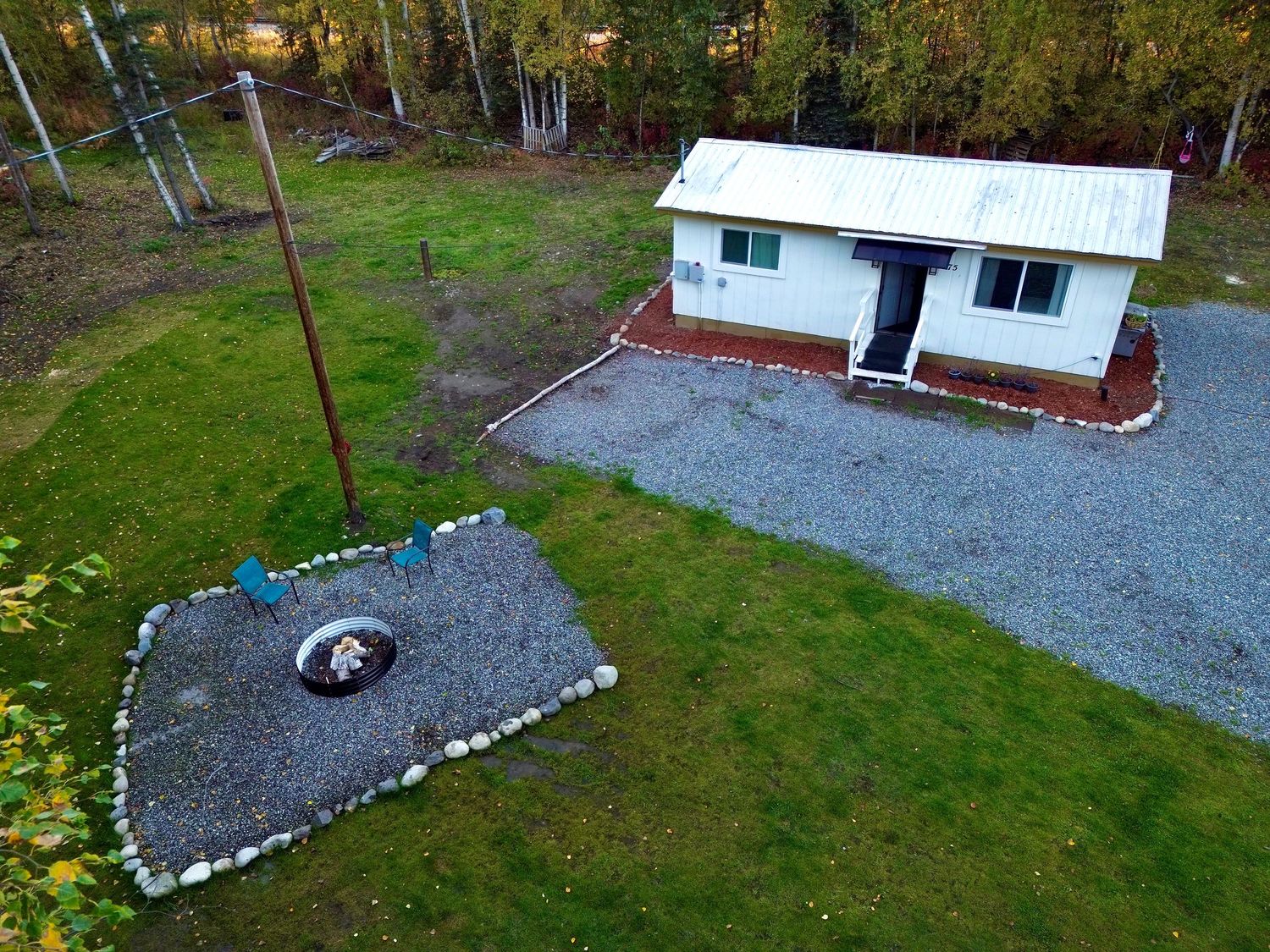 Aerial view of a small home with a white metal roof, gravel driveway, green lawn, and stone-bordered fire pit beside a wooded area.