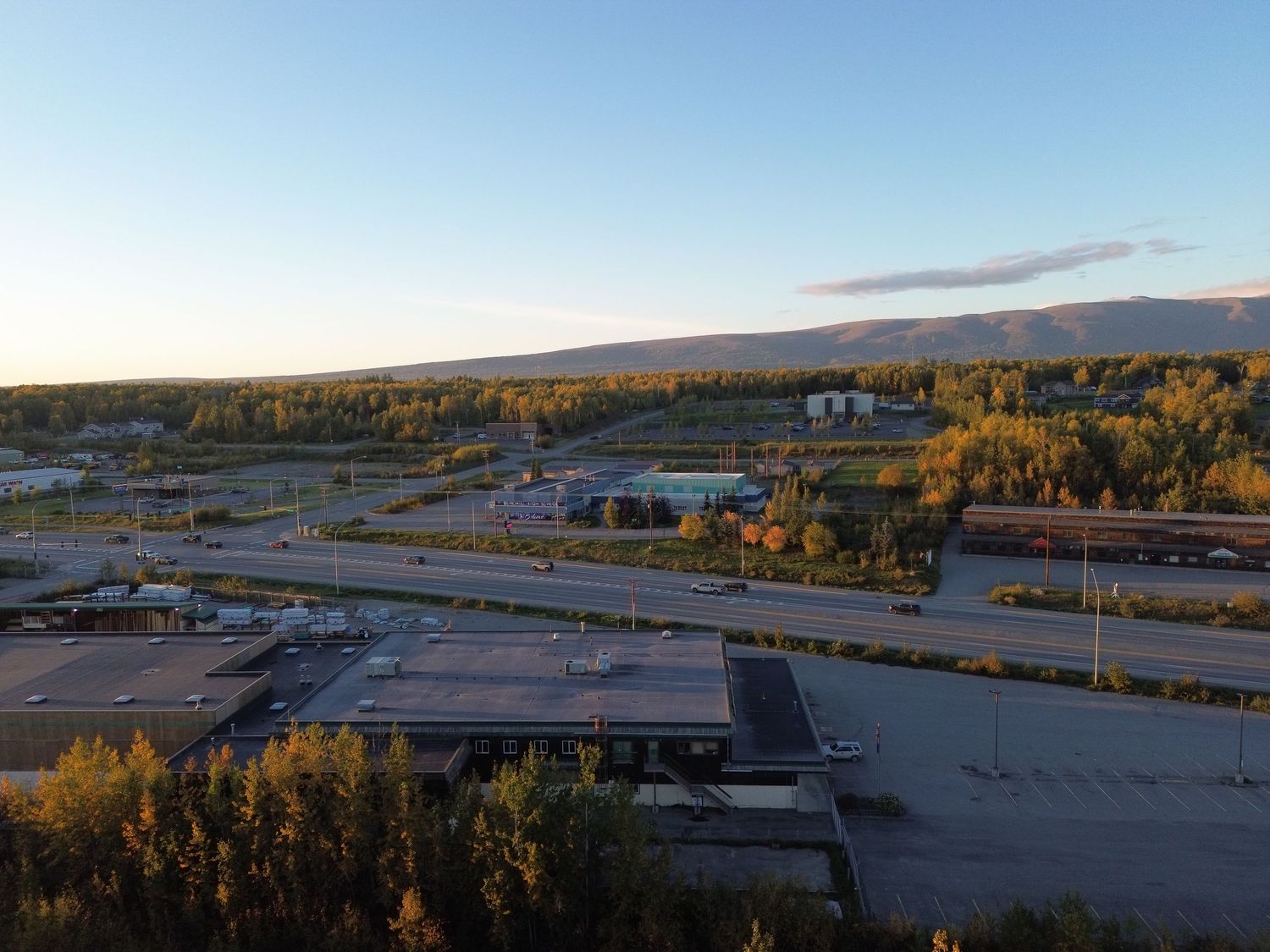Aerial view of commercial buildings and a roadway surrounded by autumn trees, with distant mountains beneath a clear evening sky.