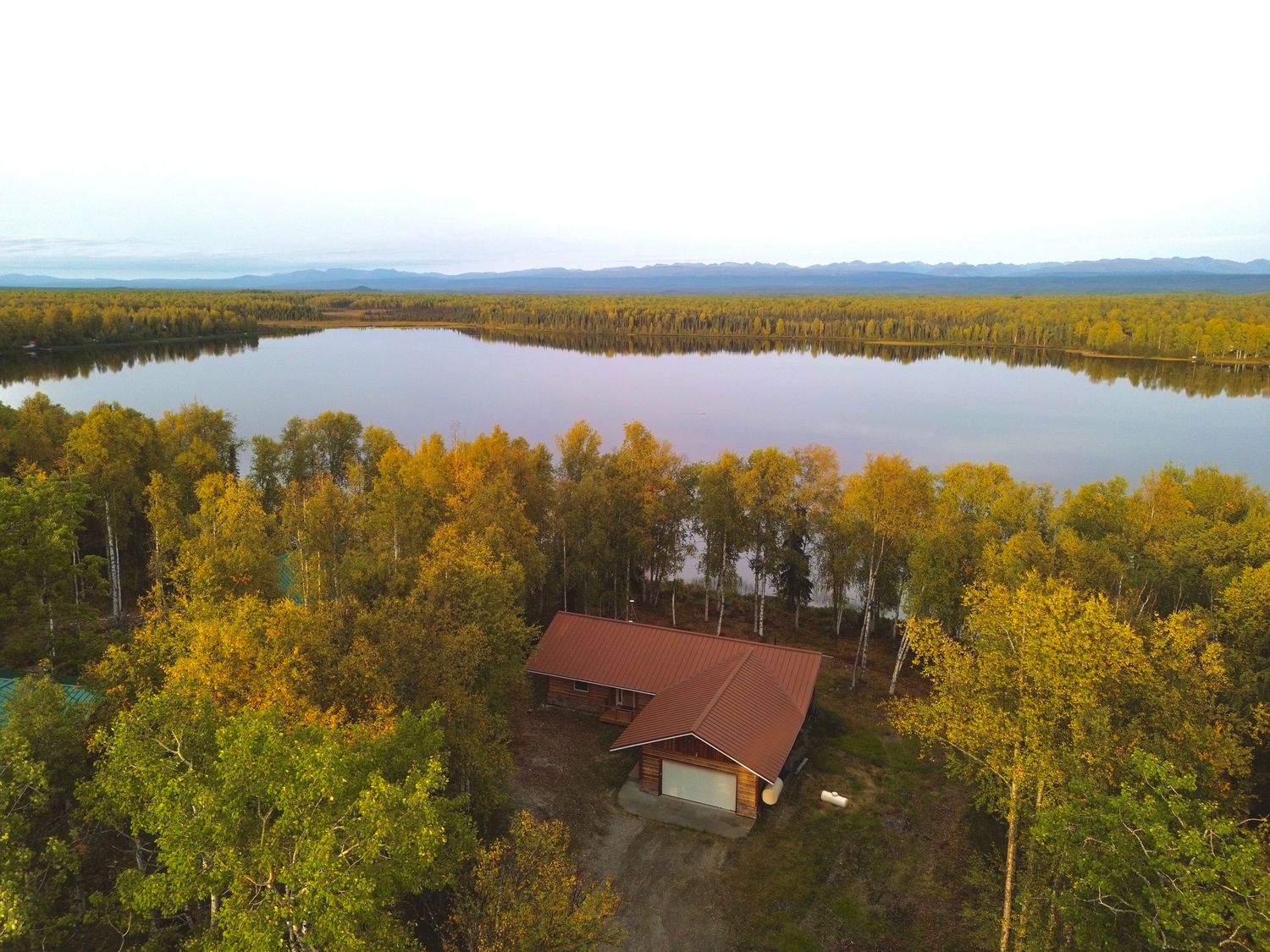 Aerial view of a home with a red roof nestled among golden autumn trees beside a lake, with forest stretching across the distant shoreline.