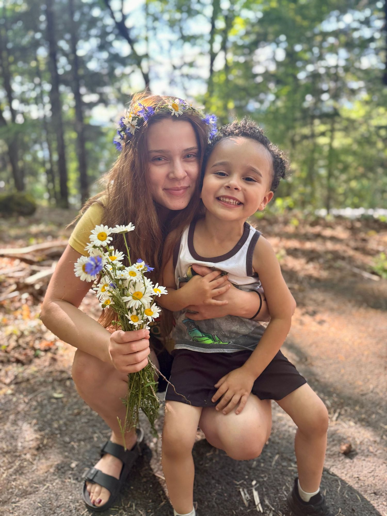 Young woman with flower crown hugging smiling toddler boy in forest.