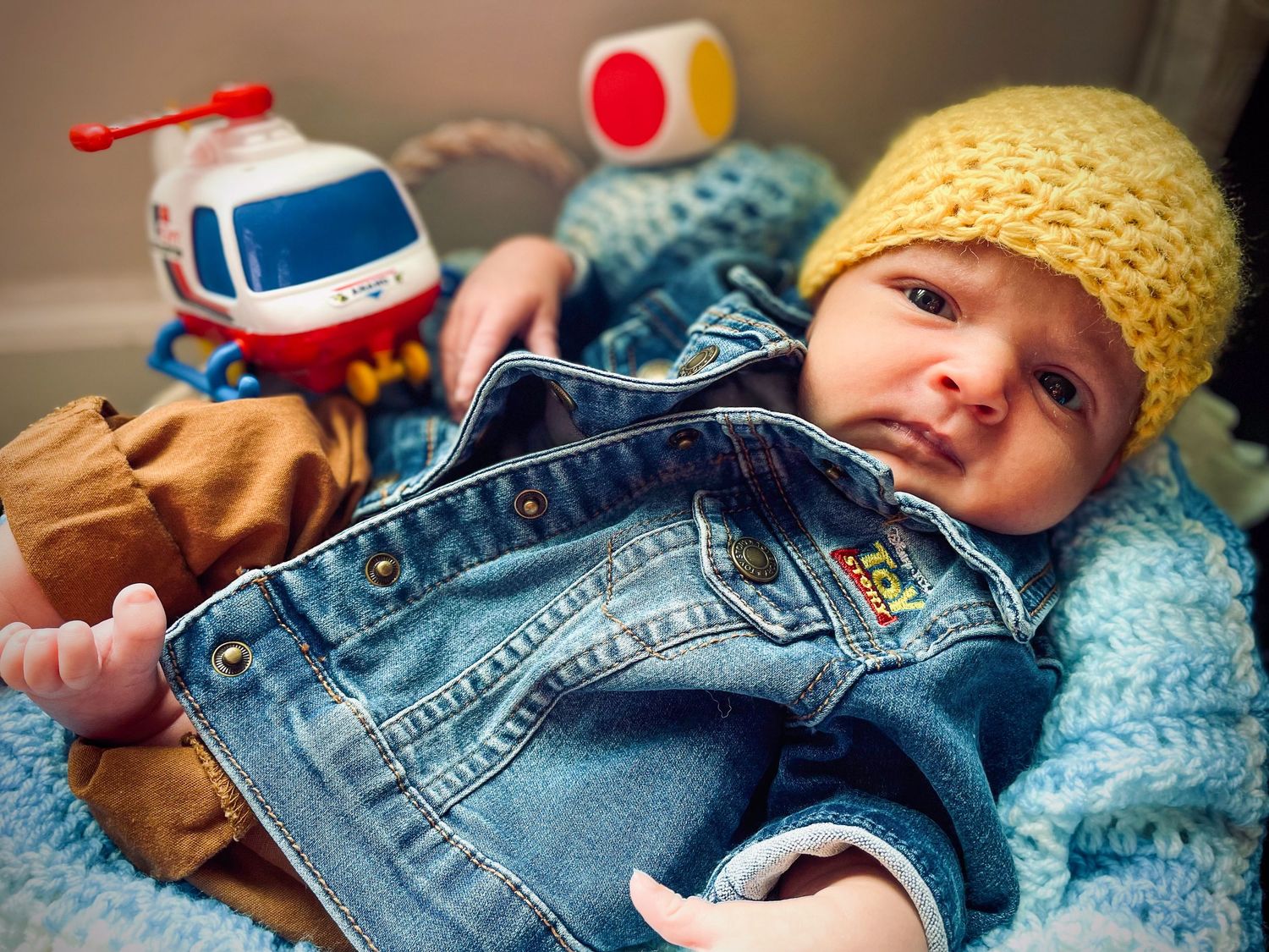 Newborn baby in denim jacket and yellow knit hat lying near colorful toys.
