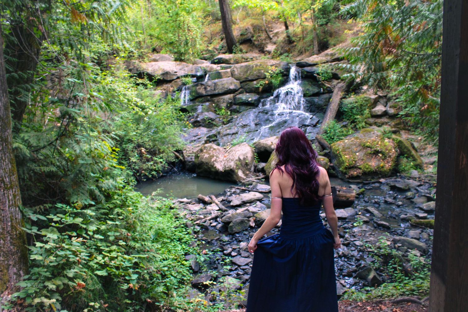 Woman in navy blue gown views scenic waterfall surrounded by lush forest greenery.
