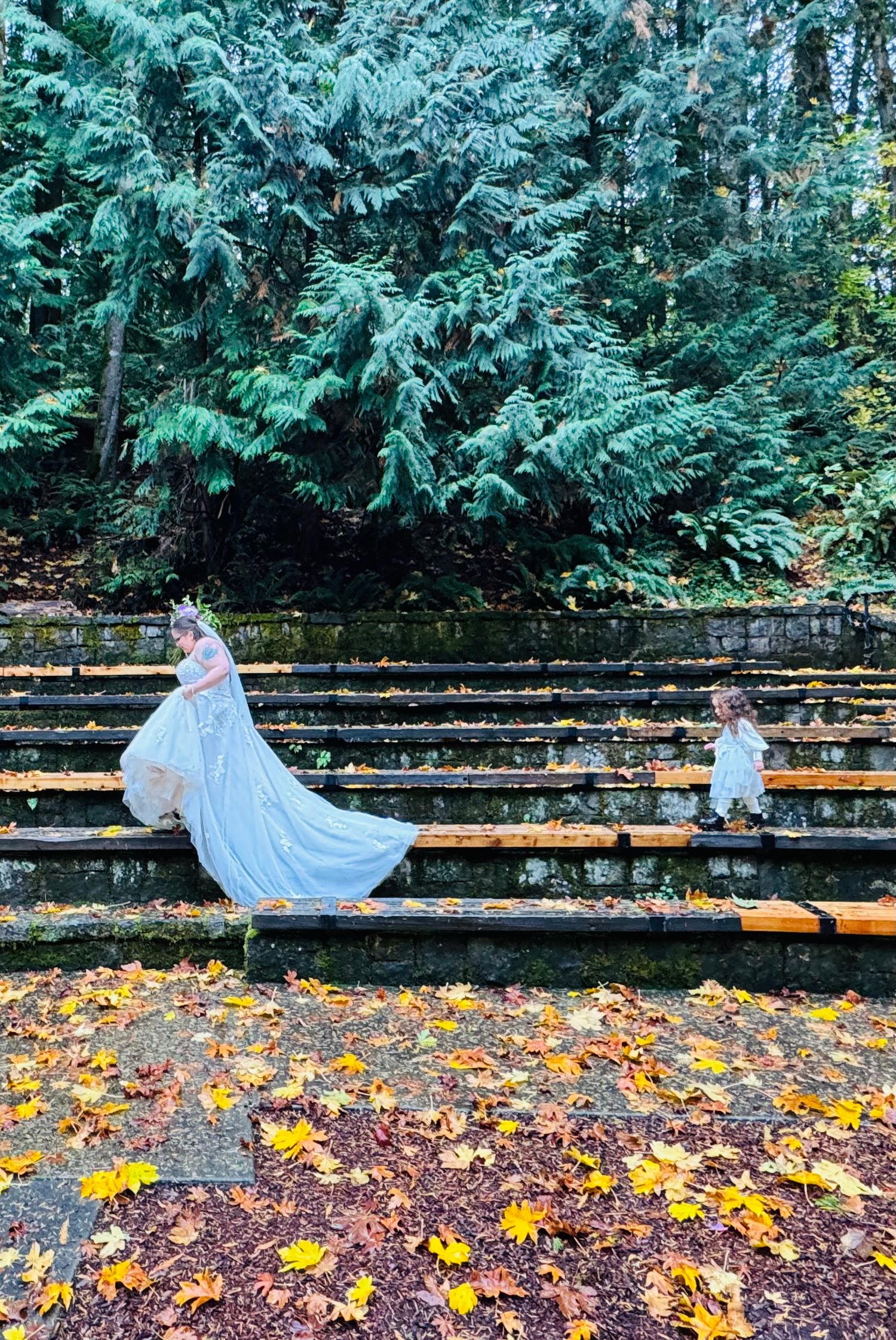 A bride in a white gown with long train poses on stone amphitheater steps surrounded by autumn leaves and evergreen trees.