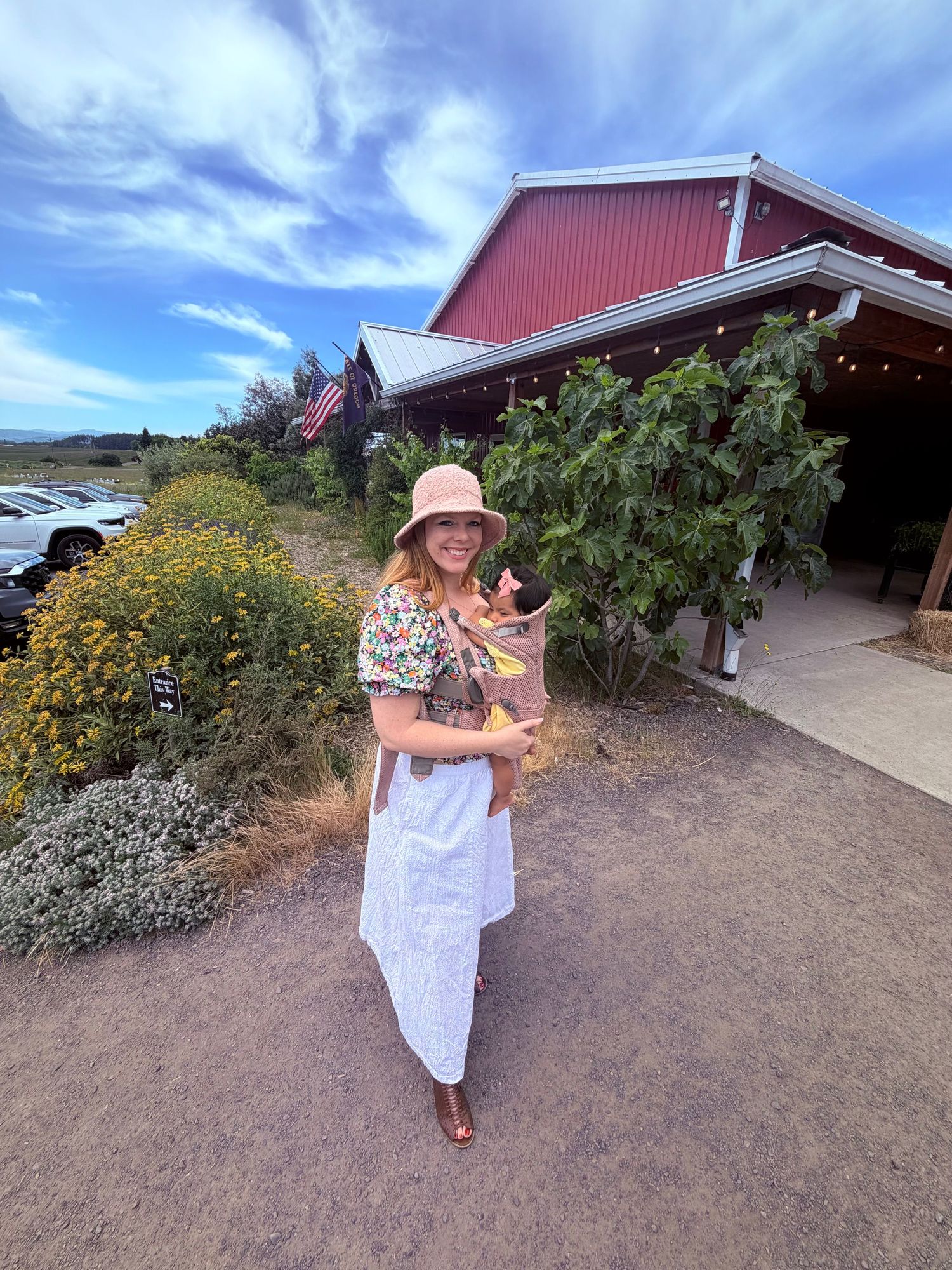 Woman in floral top and white skirt holding a baby outdoors near a red barn on a sunny day.