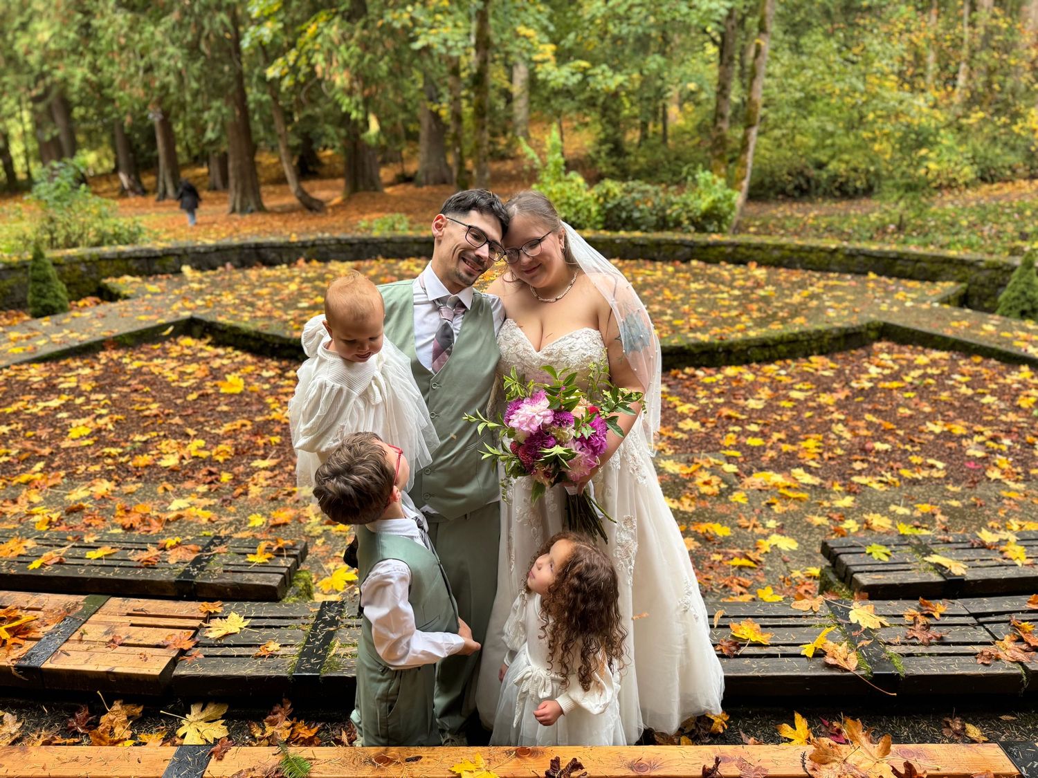 Bride and groom with two young children on autumn leaf-covered fountain.