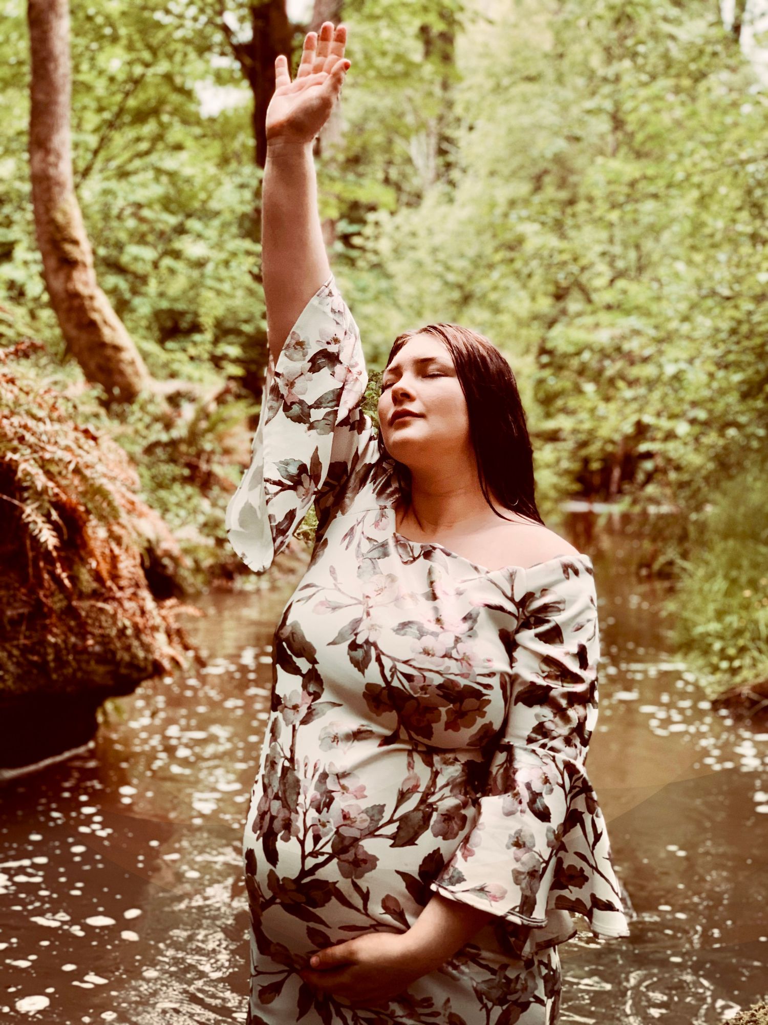 Pregnant woman in floral off-shoulder dress standing in a forest stream with one hand raised upward.