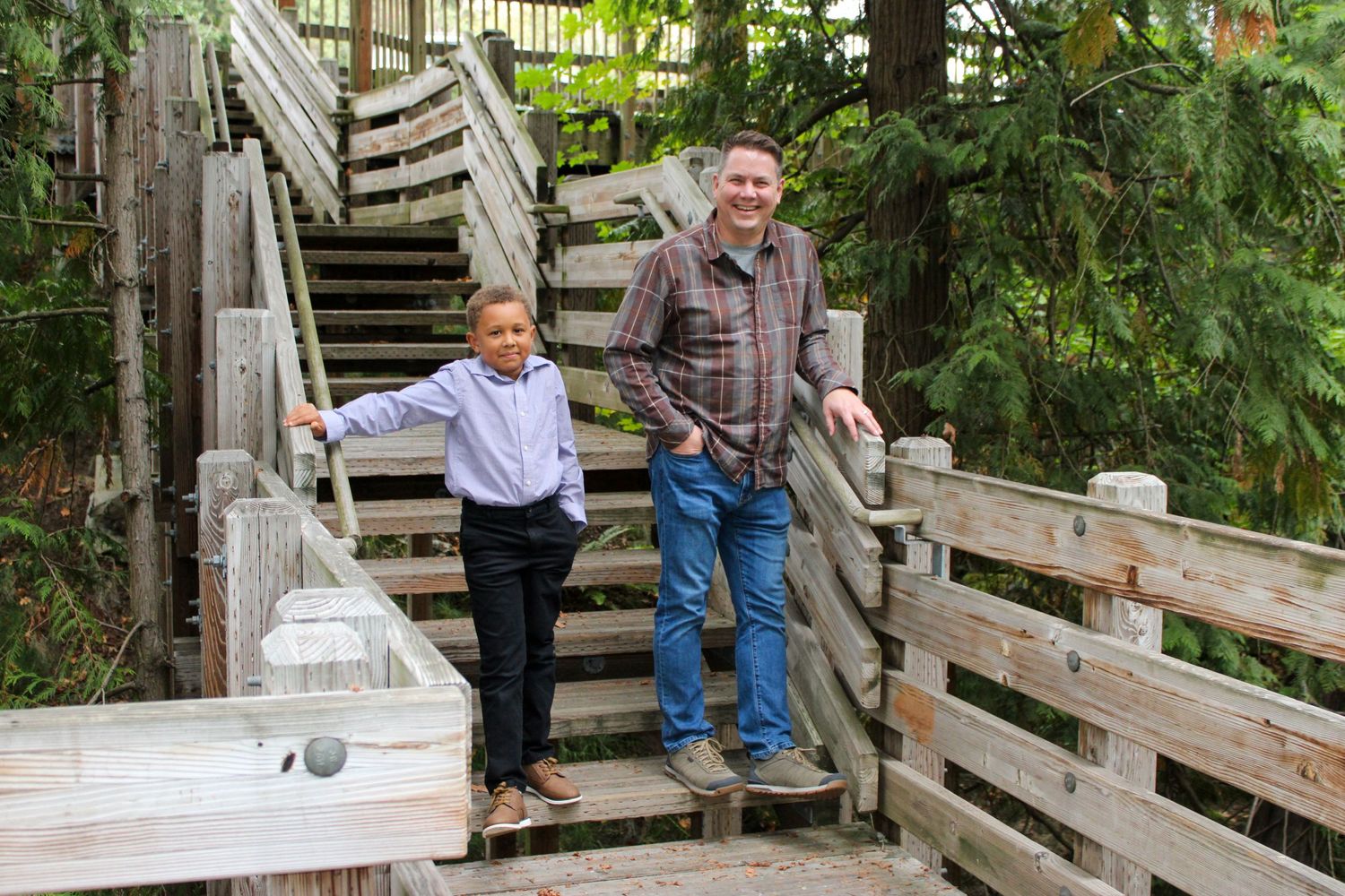 Man in plaid shirt and young boy pose on wooden outdoor staircase in forest park.