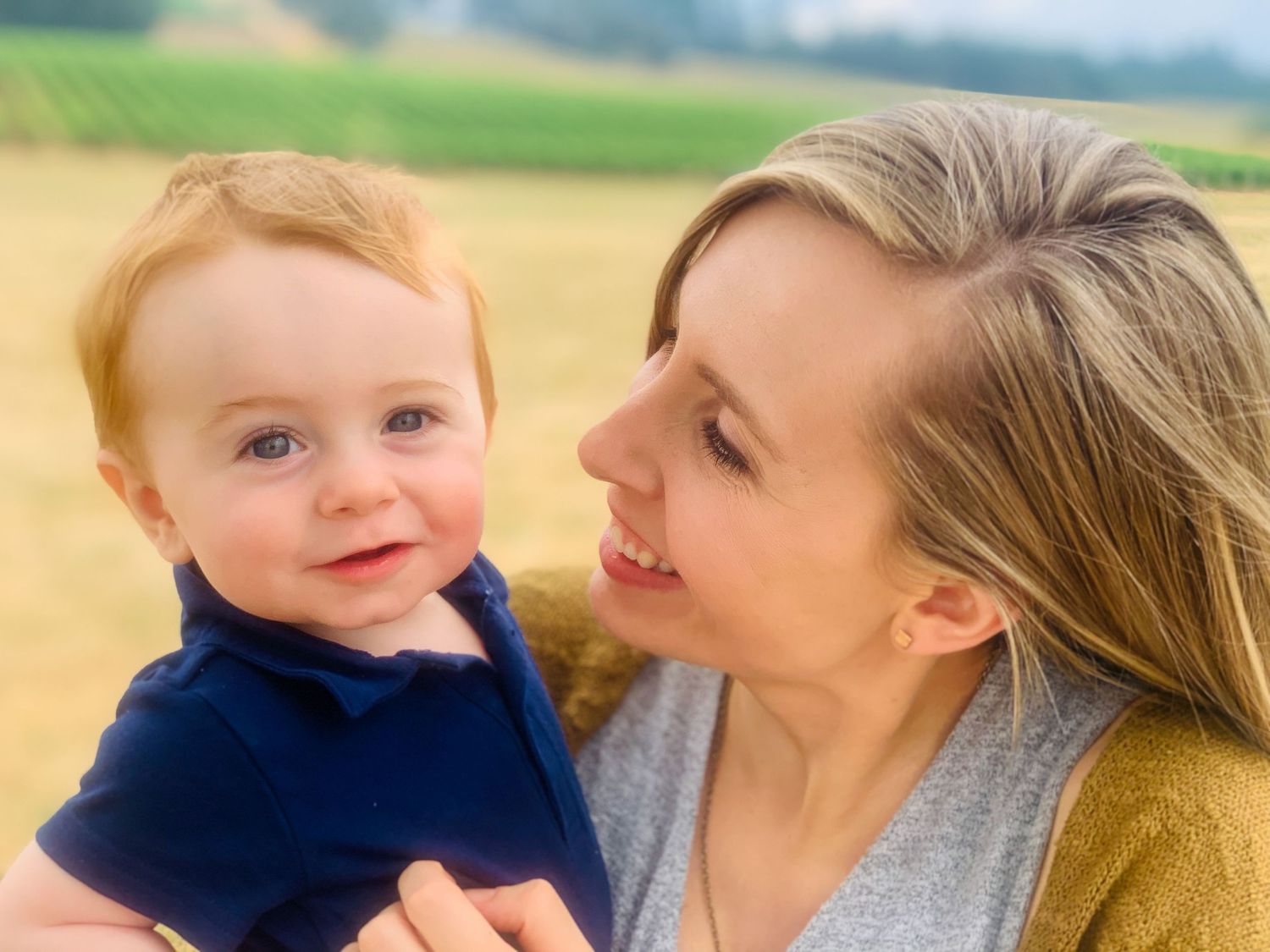 Mother gazing lovingly at her smiling red-haired baby boy in an outdoor field.
