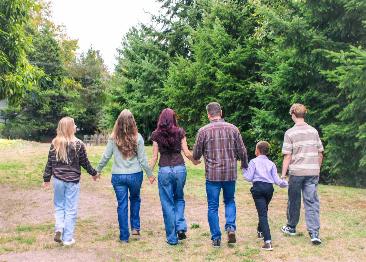 A family of six walks hand-in-hand away from the camera along a grassy path surrounded by tall green trees.