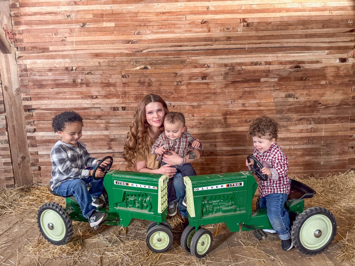 Woman with three young children posing on green pedal tractors in a rustic barn.