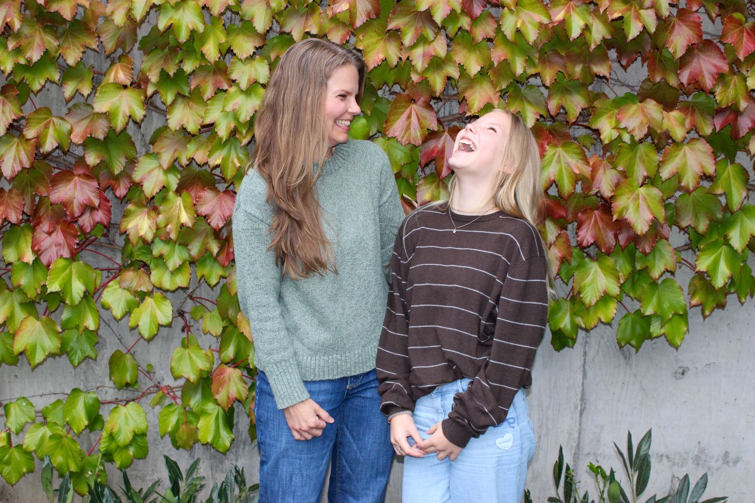 Two women in casual sweaters laugh together in front of colorful red and green ivy wall.