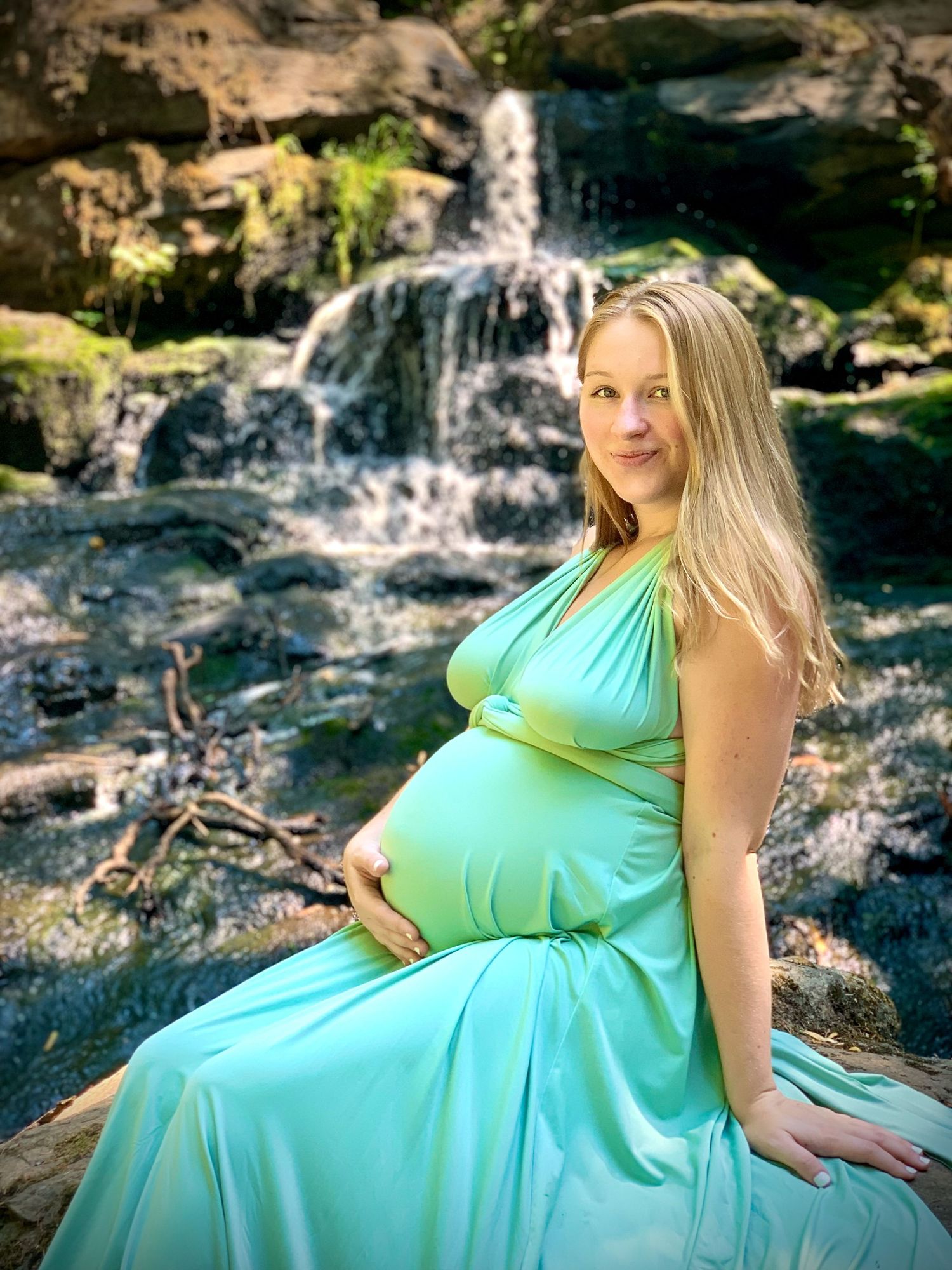 Pregnant woman in teal maternity gown sitting by a rocky waterfall outdoors.