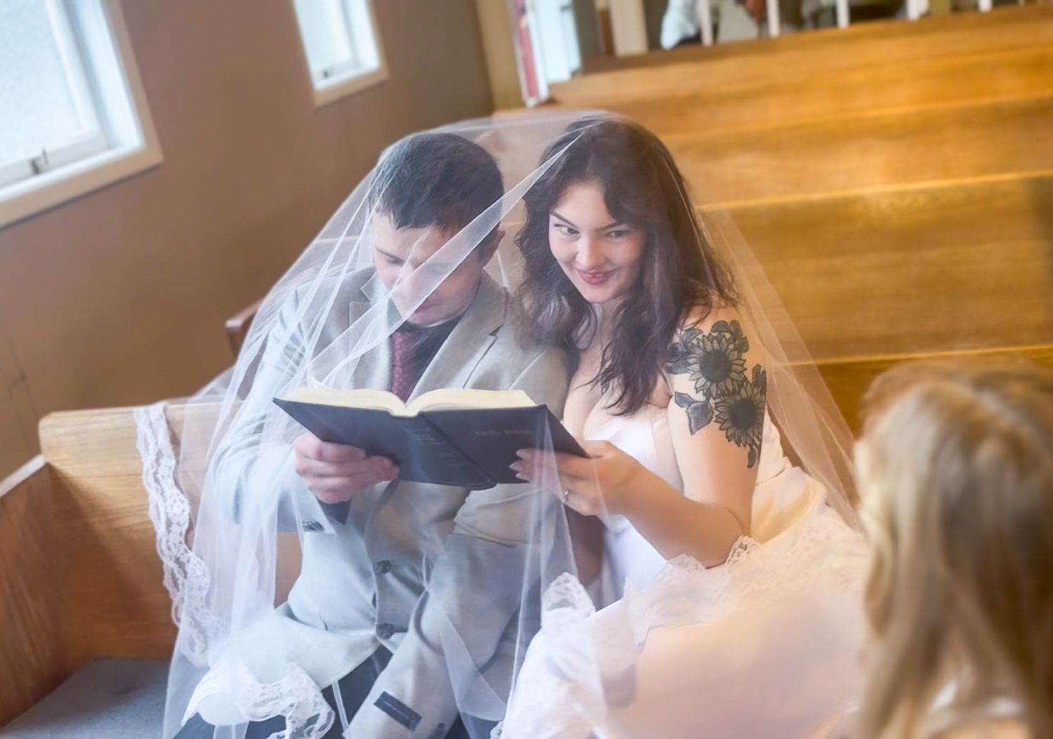 A bride and groom share an intimate moment reading a book together under a wedding veil in a cozy church.