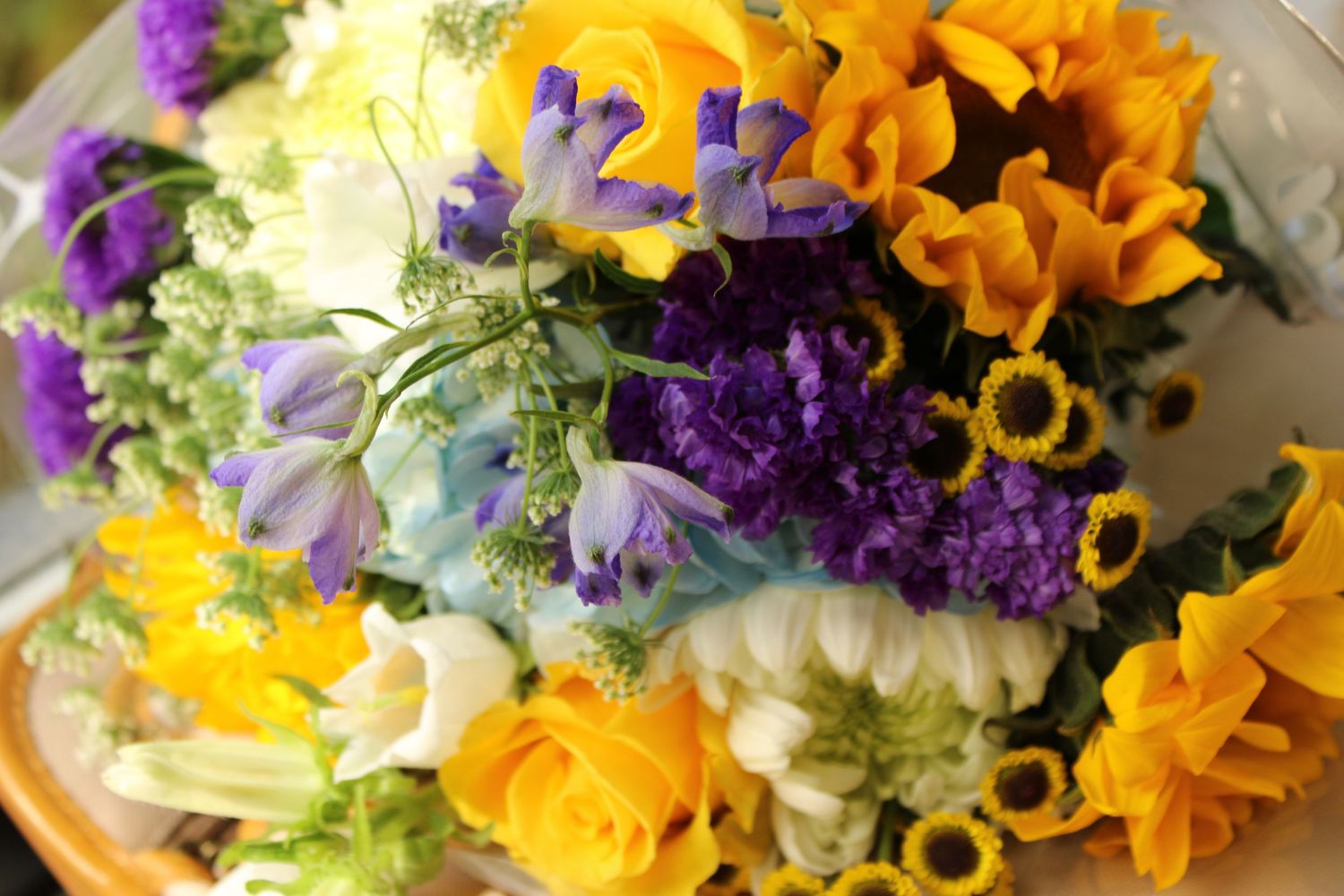 Close-up of a vibrant bouquet featuring yellow roses, purple statice, blue flowers, and sunflower accents.