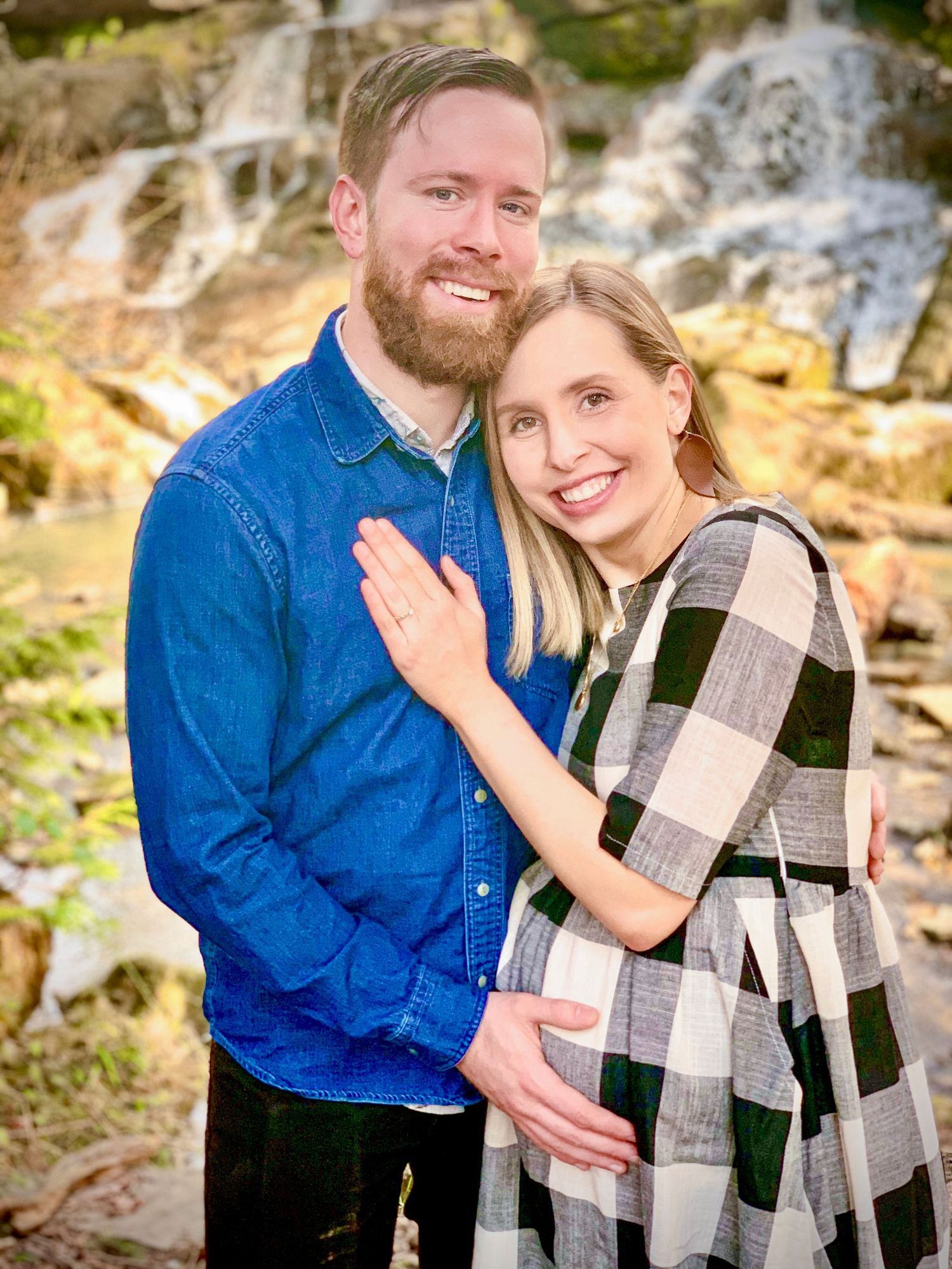 Happy expecting couple embracing in front of a waterfall, woman showing baby bump in plaid dress.
