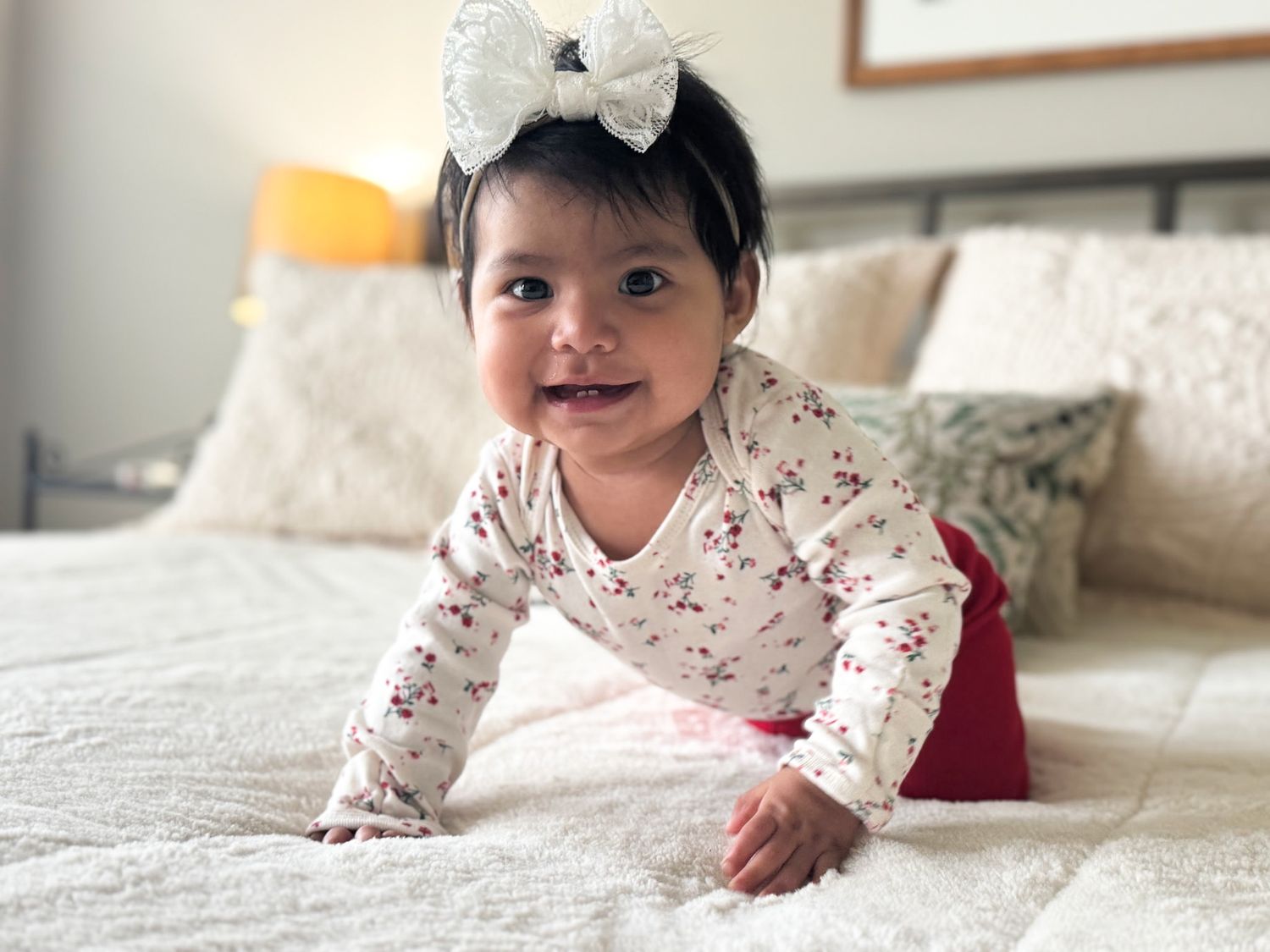 Smiling baby girl in floral onesie with white bow headband crawling on a white bed.