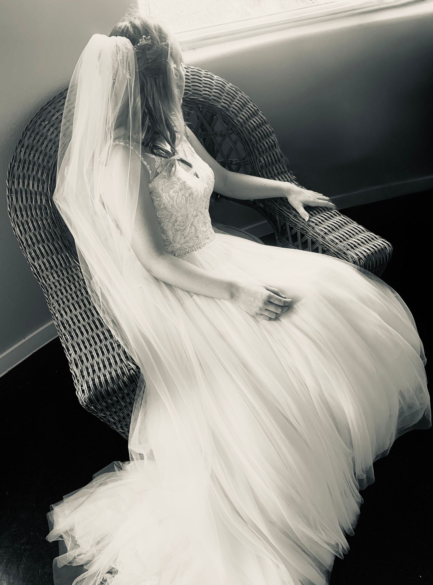 Elegant bride in a flowing white wedding gown and veil seated in a wicker chair, black and white portrait photography.