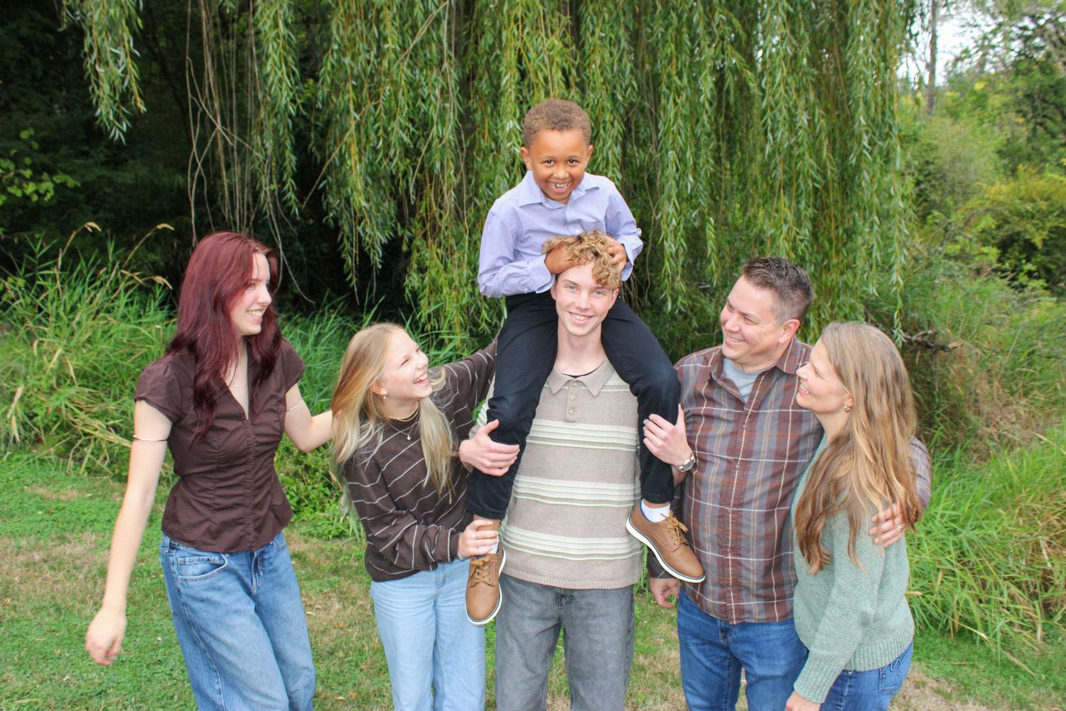 A happy family laughs outdoors under a weeping willow tree on a sunny day.
