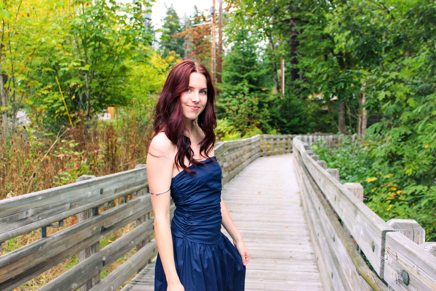 Young woman in navy blue strapless gown poses on wooden bridge surrounded by lush green trees.